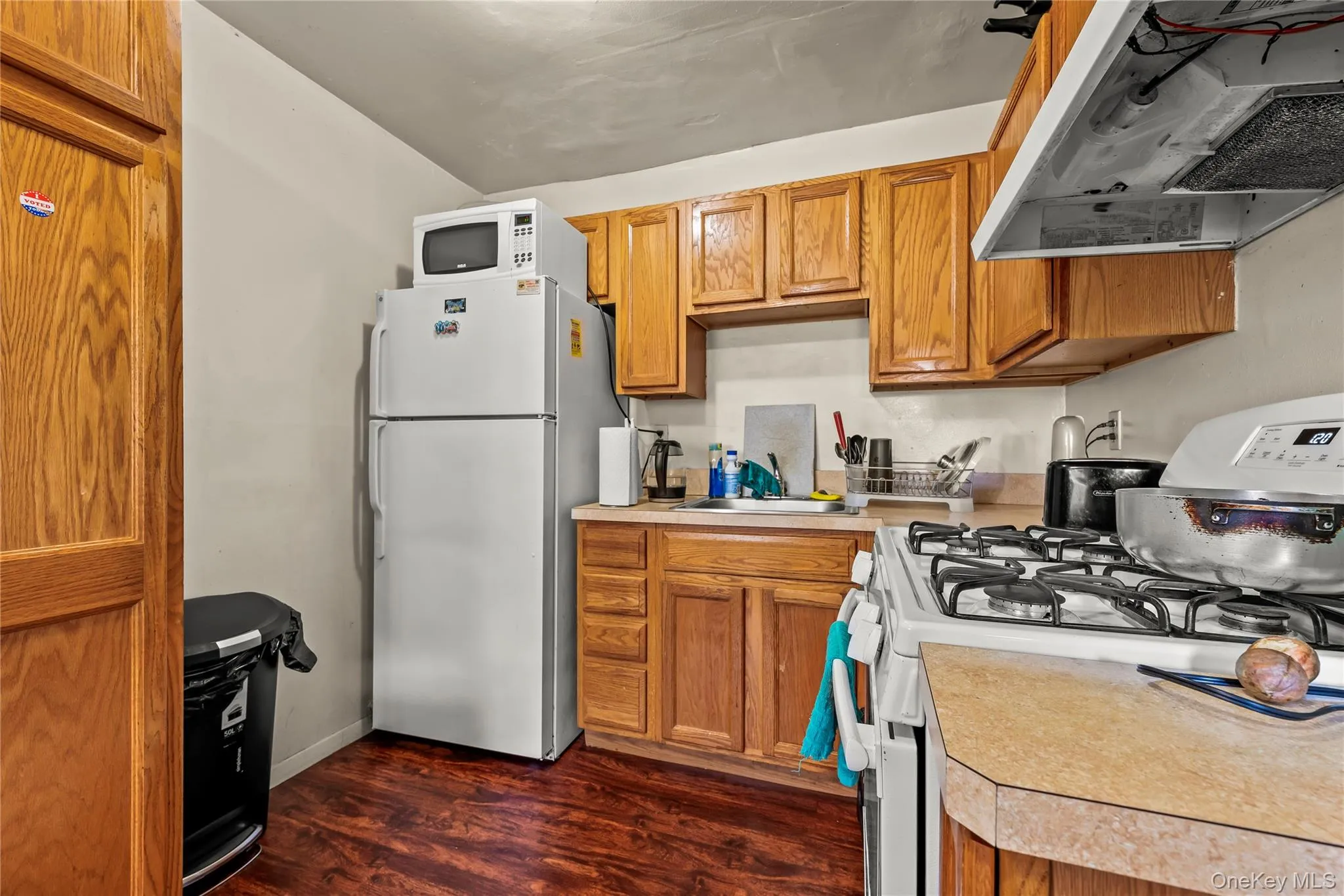 Kitchen featuring white appliances, wall chimney range hood, light countertops, dark wood-style floors, and brown cabinets Kitchen featuring white appliances, wall chimney range hood, light countertops, dark wood-style floors, and brown cabinets