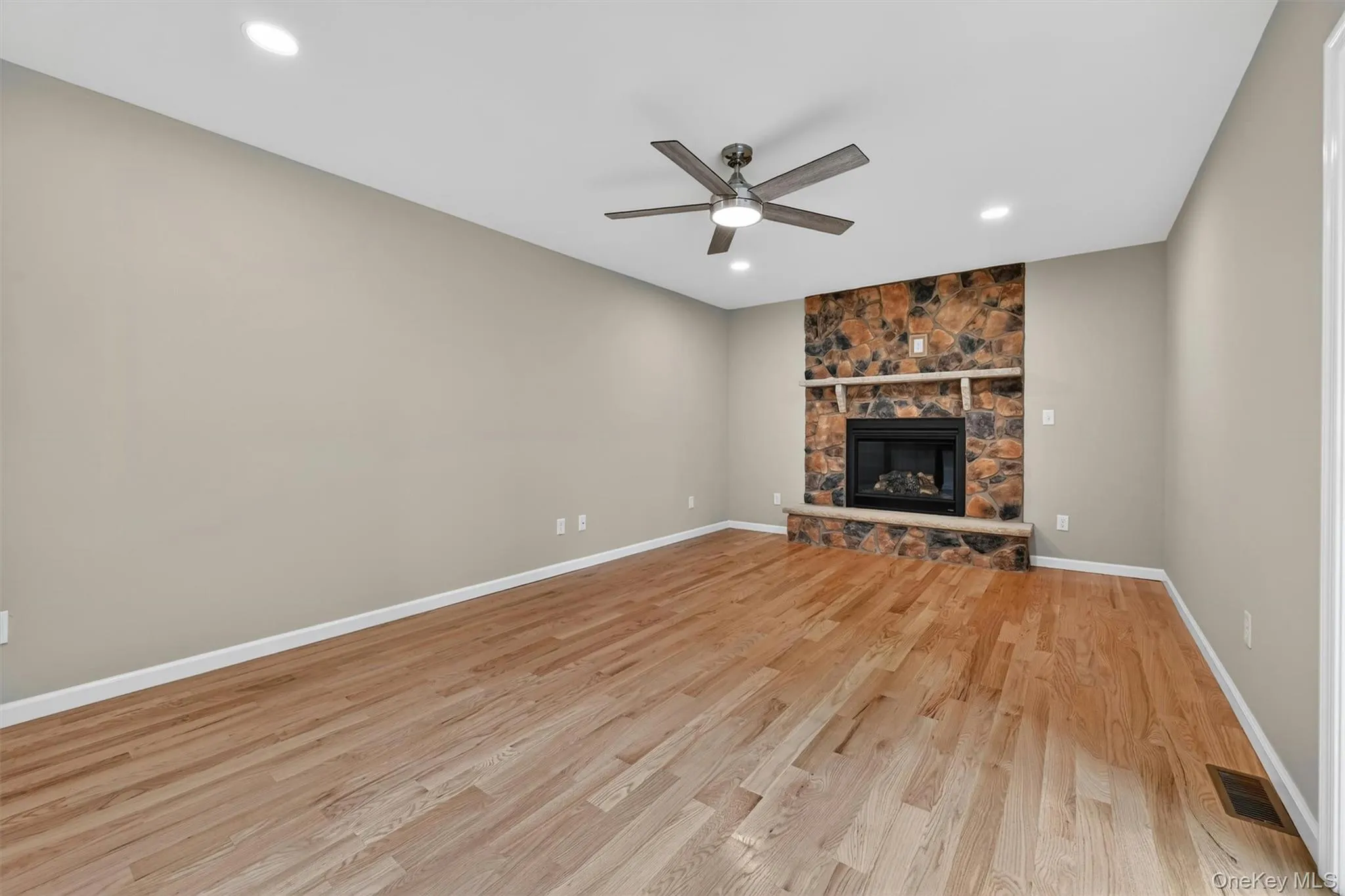 Spacious room featuring a stone-clad fireplace with a hearth, wood-finish flooring, a ceiling fan, and recessed lighting Spacious room featuring a stone-clad fireplace with a hearth, wood-finish flooring, a ceiling fan, and recessed lighting