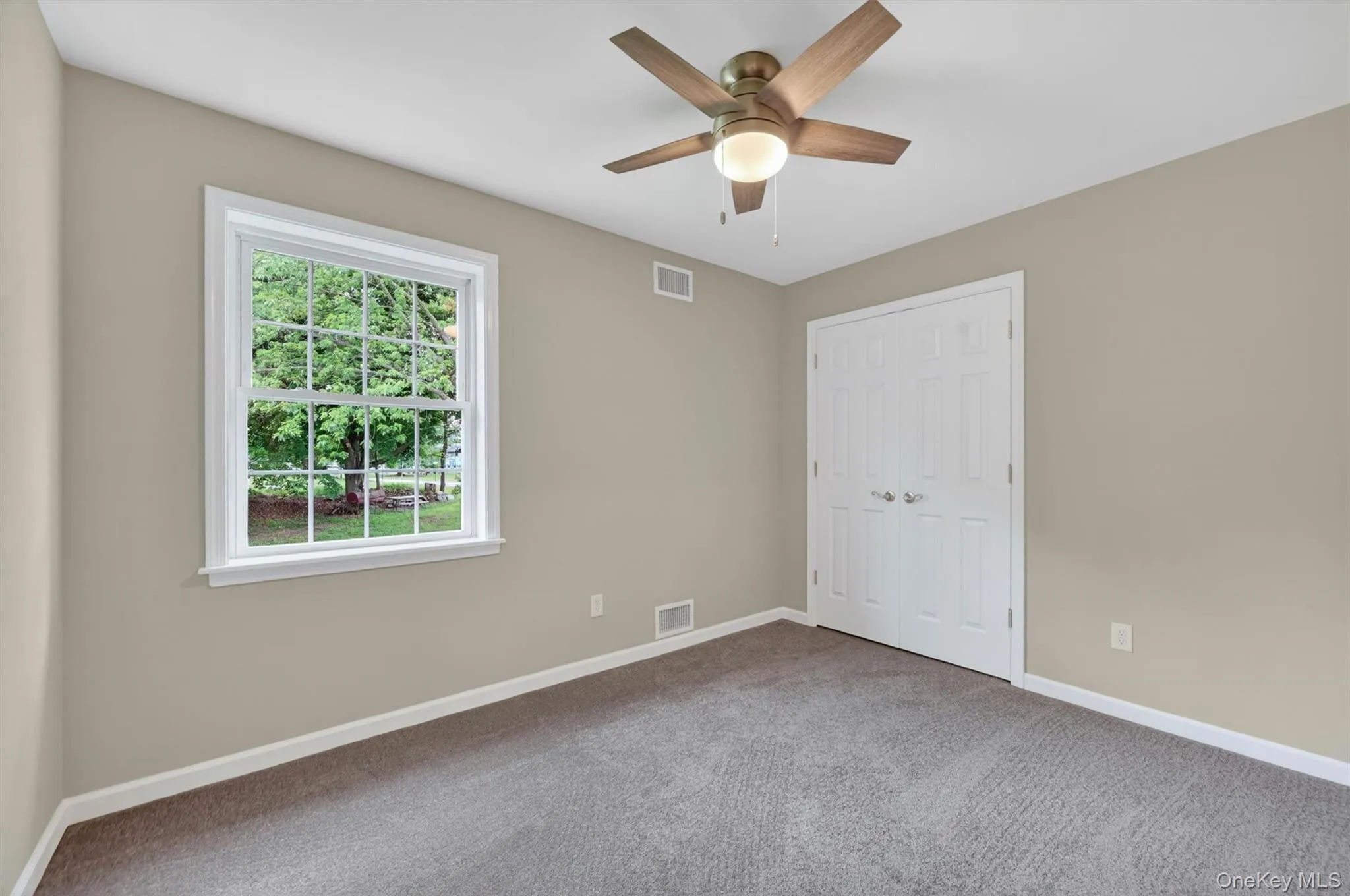 Bedroom featuring neutral wall paint, gray carpet, a white framed window with outdoor greenery, a ceiling fan with light fixture, and white bi-fold closet doors Bedroom featuring neutral wall paint, gray carpet, a white framed window with outdoor greenery, a ceiling fan with light fixture, and white bi-fold closet doors
