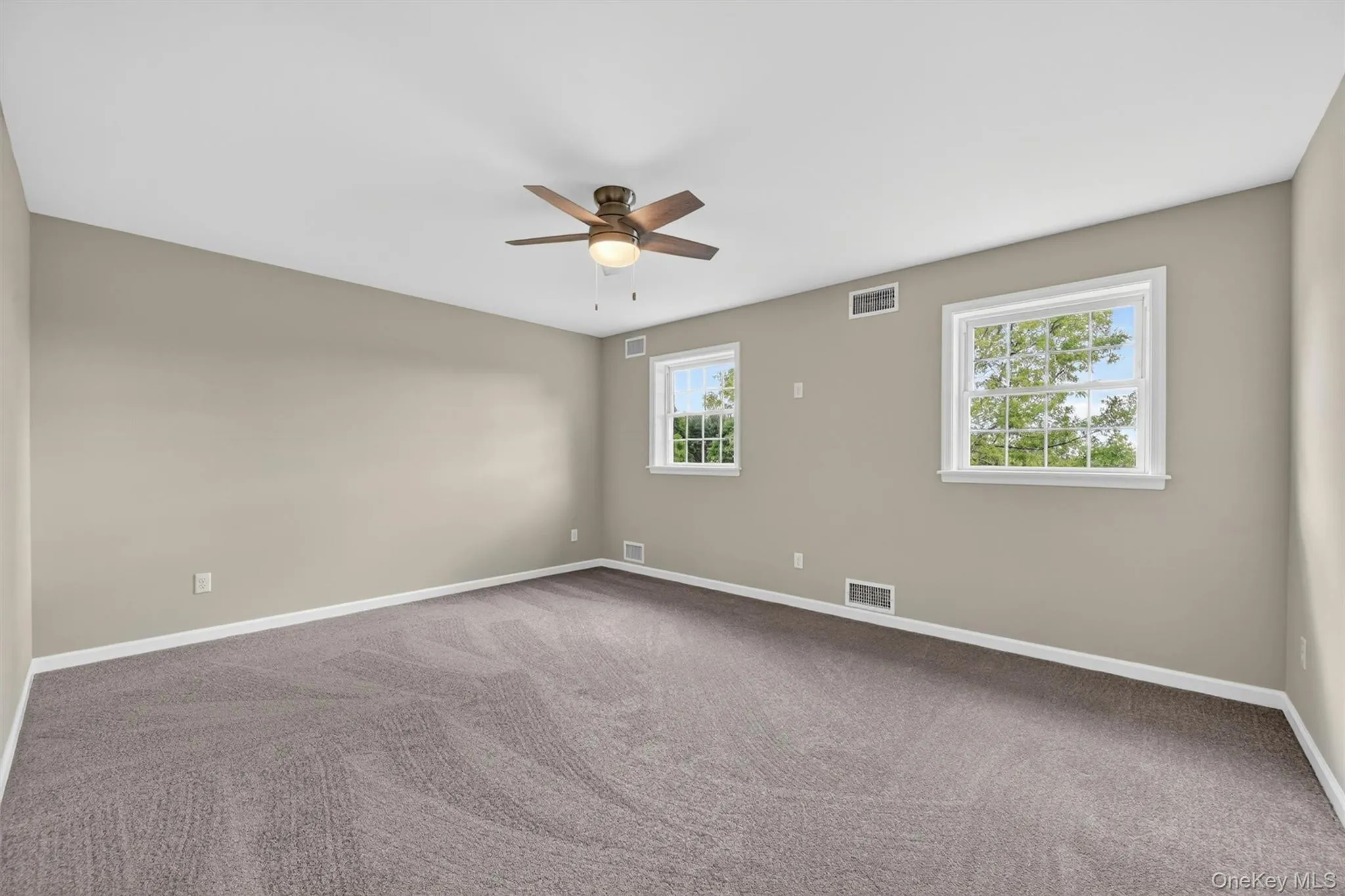 Spacious room featuring neutral-toned walls, gray carpet, two windows with white trim, a ceiling fan with integrated lighting, and white baseboards Spacious room featuring neutral-toned walls, gray carpet, two windows with white trim, a ceiling fan with integrated lighting, and white baseboards