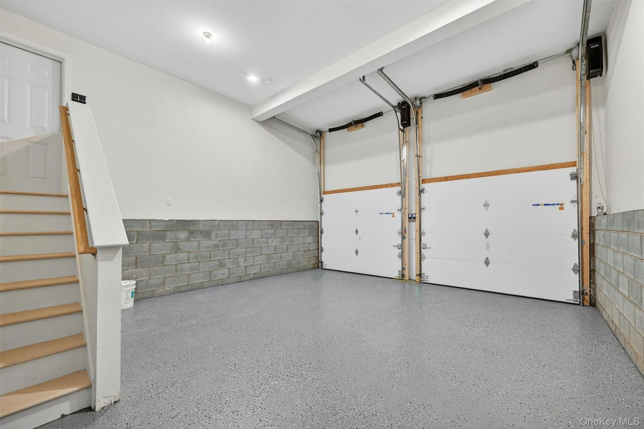 Two-car garage featuring a speckled epoxy floor coating, two white panel garage doors, concrete block wainscoting, and recessed lighting Two-car garage featuring a speckled epoxy floor coating, two white panel garage doors, concrete block wainscoting, and recessed lighting
