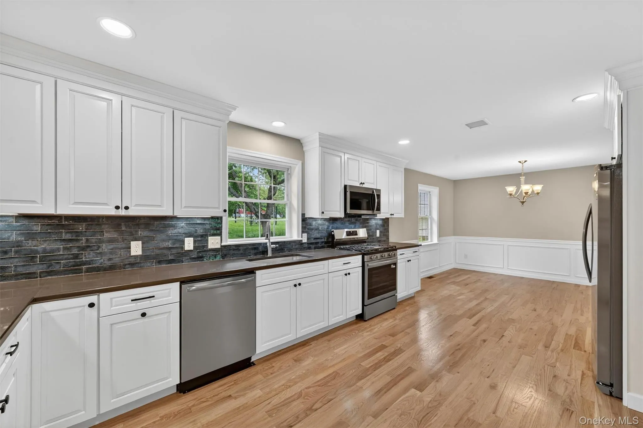 Kitchen featuring white shaker-style cabinetry, stainless steel appliances, dark subway tile backsplash, and light wood-finish flooring Kitchen featuring white shaker-style cabinetry, stainless steel appliances, dark subway tile backsplash, and light wood-finish flooring