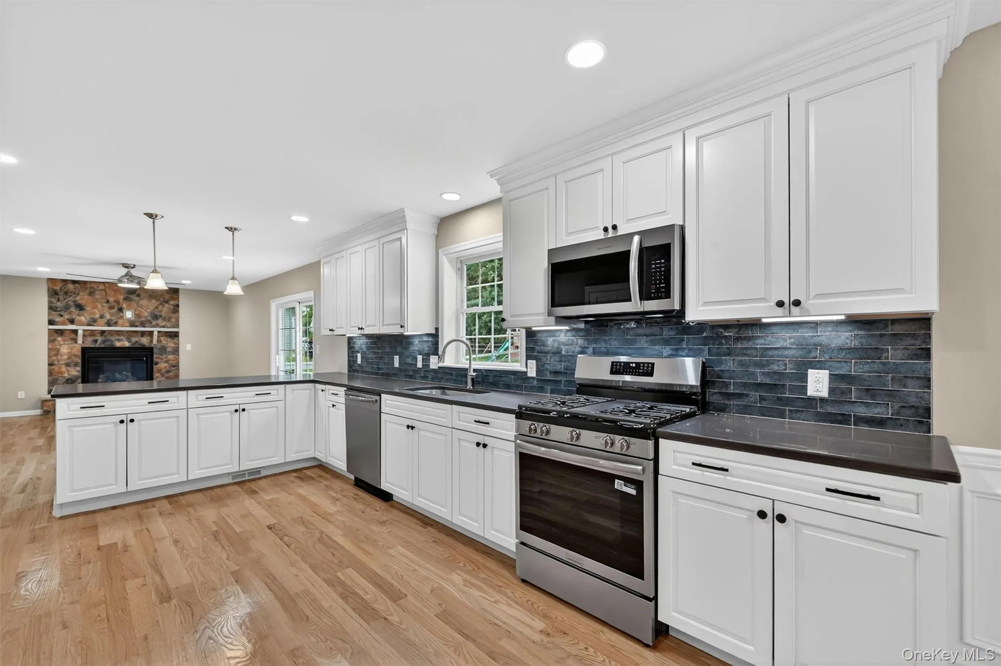 Kitchen featuring white shaker cabinetry, dark countertops, stainless steel appliances, a dark tile backsplash, and wood-finish flooring Kitchen featuring white shaker cabinetry, dark countertops, stainless steel appliances, a dark tile backsplash, and wood-finish flooring