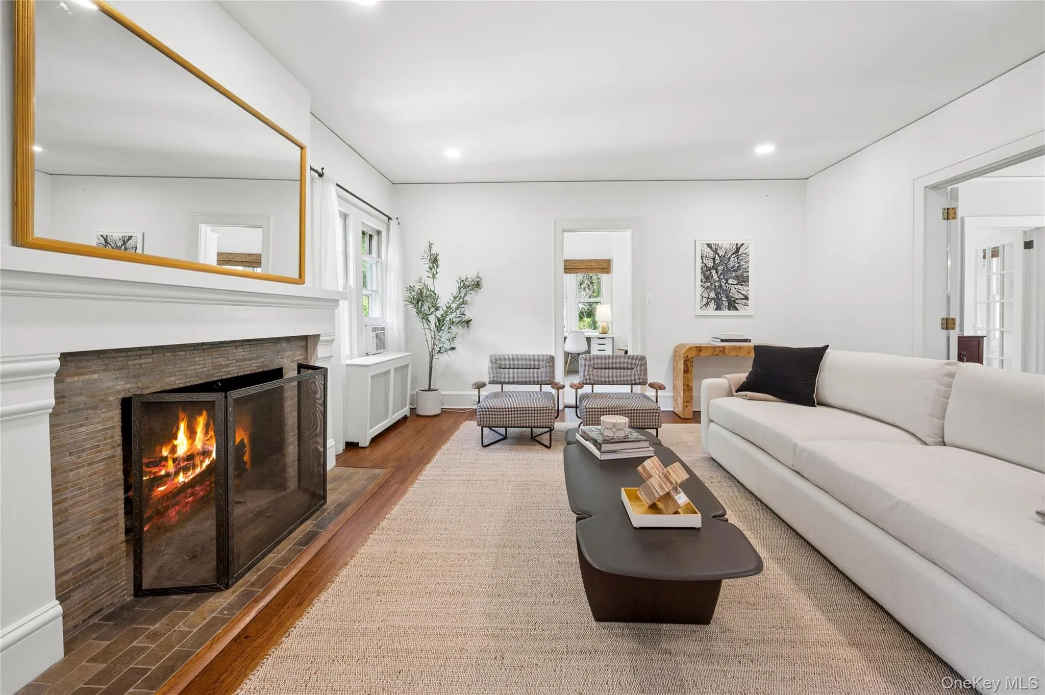 Living room featuring a stone-faced fireplace with a white mantel, hardwood flooring, recessed lighting, white walls, and an open doorway Living room featuring a stone-faced fireplace with a white mantel, hardwood flooring, recessed lighting, white walls, and an open doorway