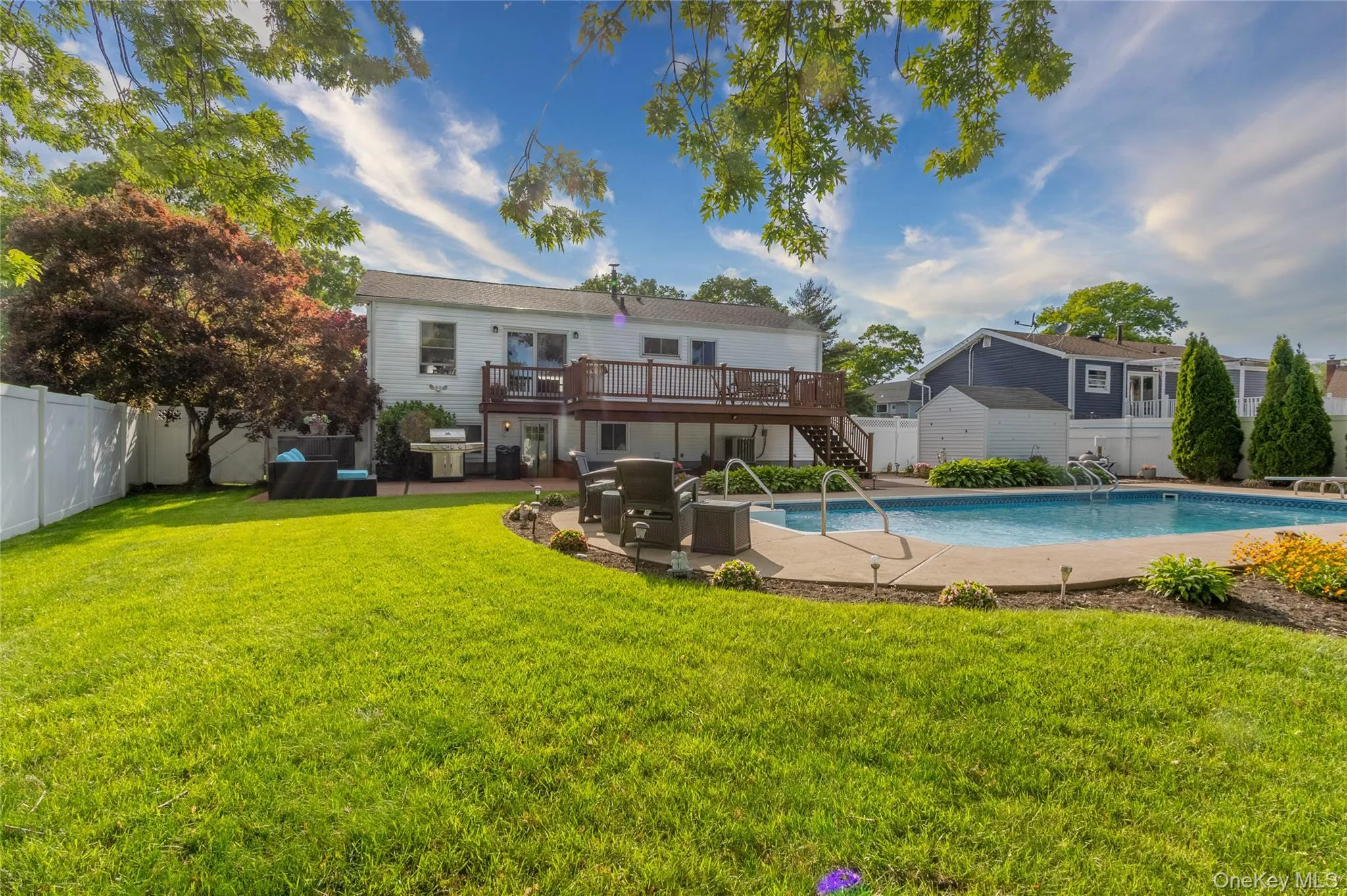 Expansive backyard featuring a large in-ground swimming pool with concrete surround, a spacious wood deck, and a lower-level patio with stone pavers Expansive backyard featuring a large in-ground swimming pool with concrete surround, a spacious wood deck, and a lower-level patio with stone pavers