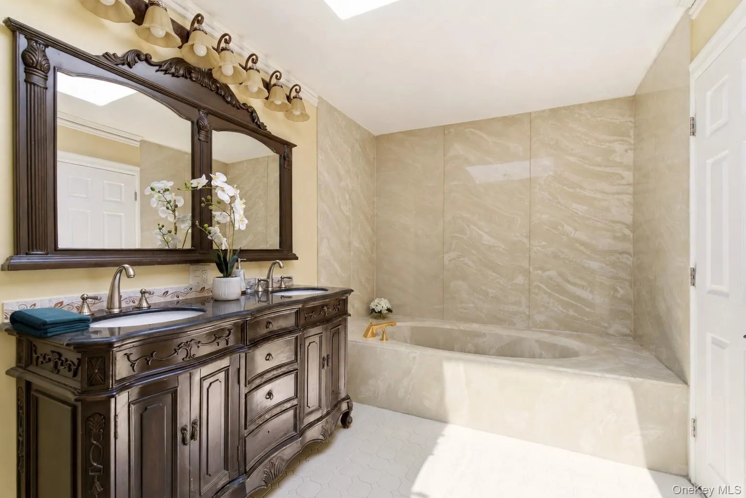 Spacious bathroom featuring a carved dual vanity with undermount sinks, a large framed mirror, and an integrated soaking tub with stone-look surround Spacious bathroom featuring a carved dual vanity with undermount sinks, a large framed mirror, and an integrated soaking tub with stone-look surround
