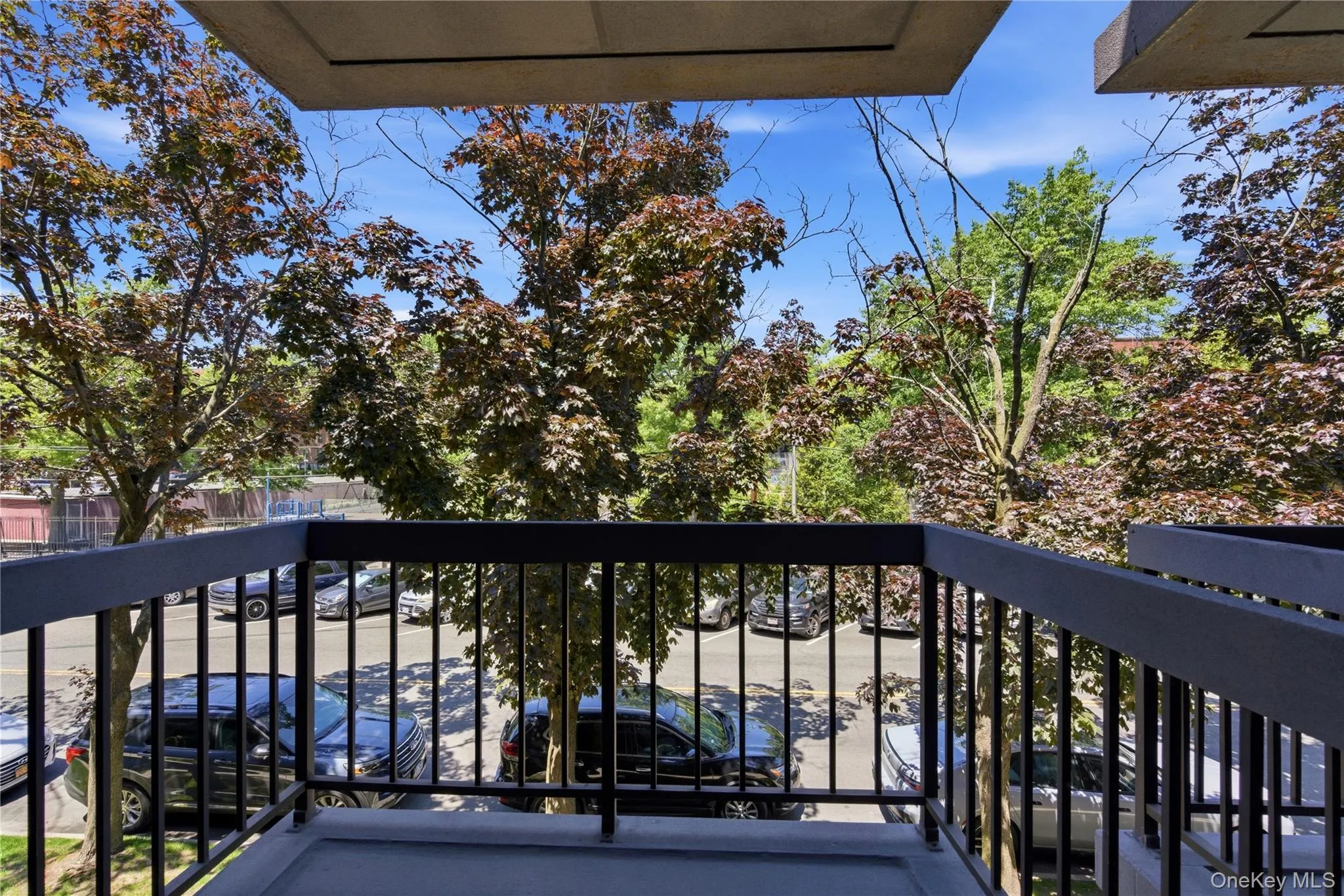 Private balcony with dark gray railing and concrete flooring, offering an elevated perspective of the surrounding mature foliage and a paved parking area Private balcony with dark gray railing and concrete flooring, offering an elevated perspective of the surrounding mature foliage and a paved parking area