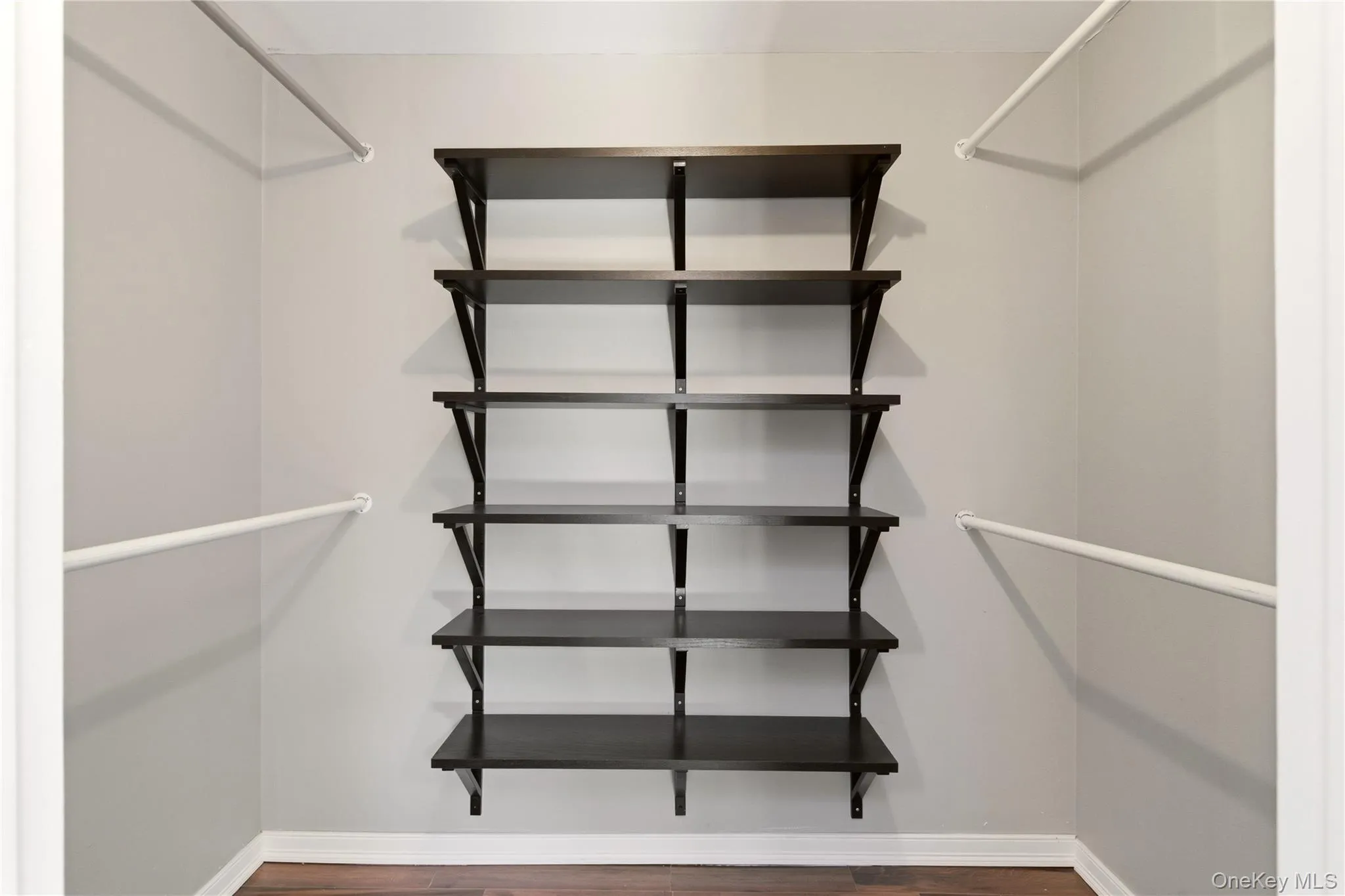 Customizable closet system featuring five dark wood-finish shelves, two hanging rods, light gray wall paint, white baseboards, and wood-finish flooring Customizable closet system featuring five dark wood-finish shelves, two hanging rods, light gray wall paint, white baseboards, and wood-finish flooring
