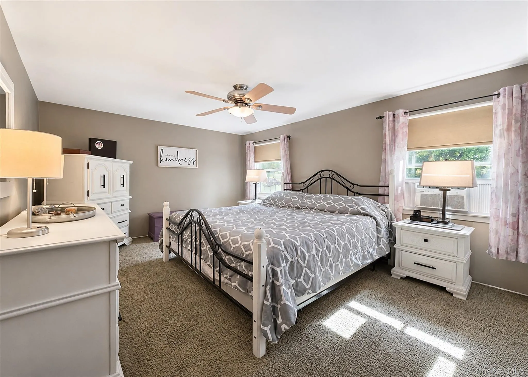 Carpeted bedroom featuring a ceiling fan with integrated lighting, two windows with roller shades, and neutral wall tones Carpeted bedroom featuring a ceiling fan with integrated lighting, two windows with roller shades, and neutral wall tones