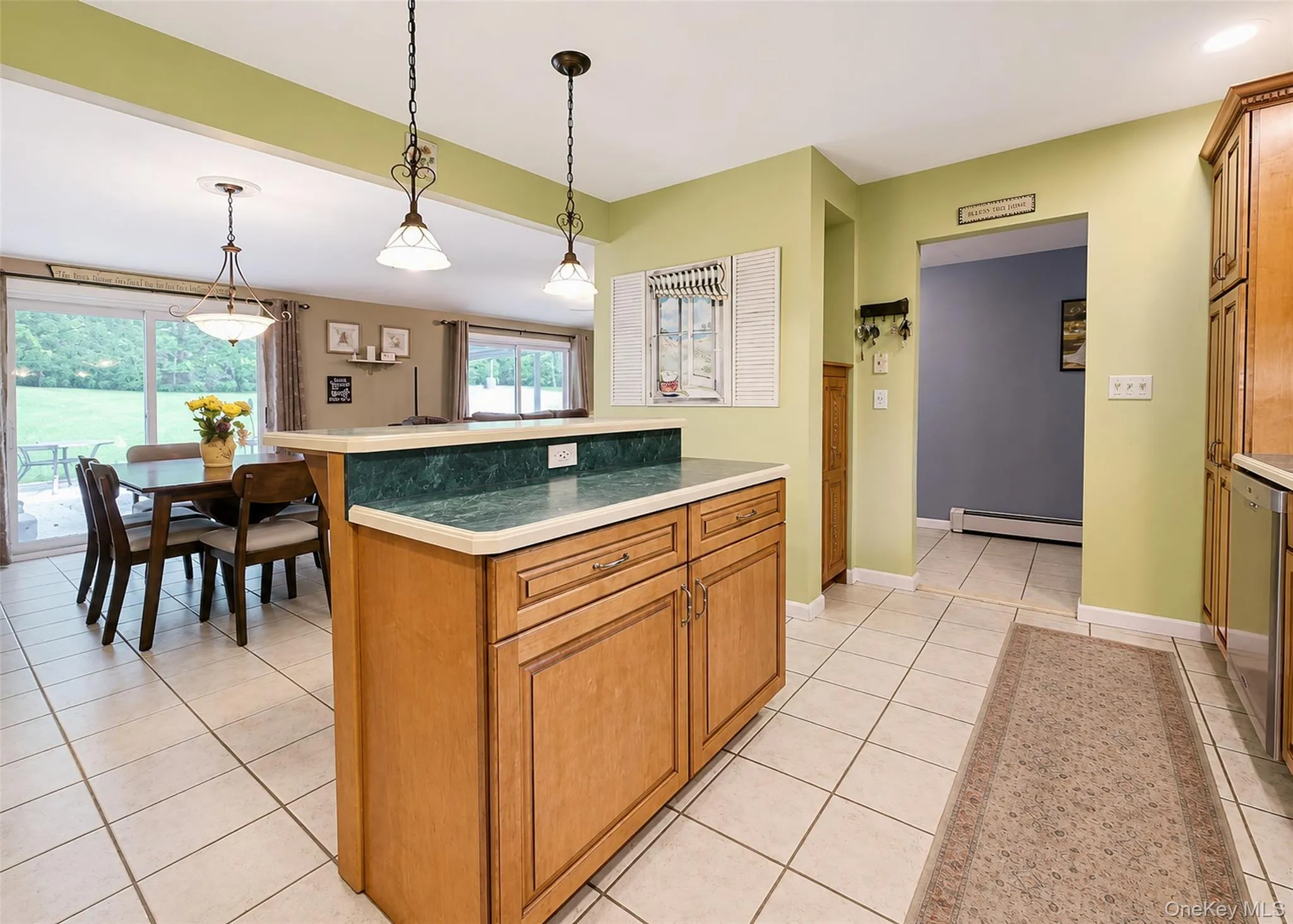 Kitchen featuring light wood cabinetry, a two-tiered island with a light countertop and green backsplash, and tile flooring Kitchen featuring light wood cabinetry, a two-tiered island with a light countertop and green backsplash, and tile flooring