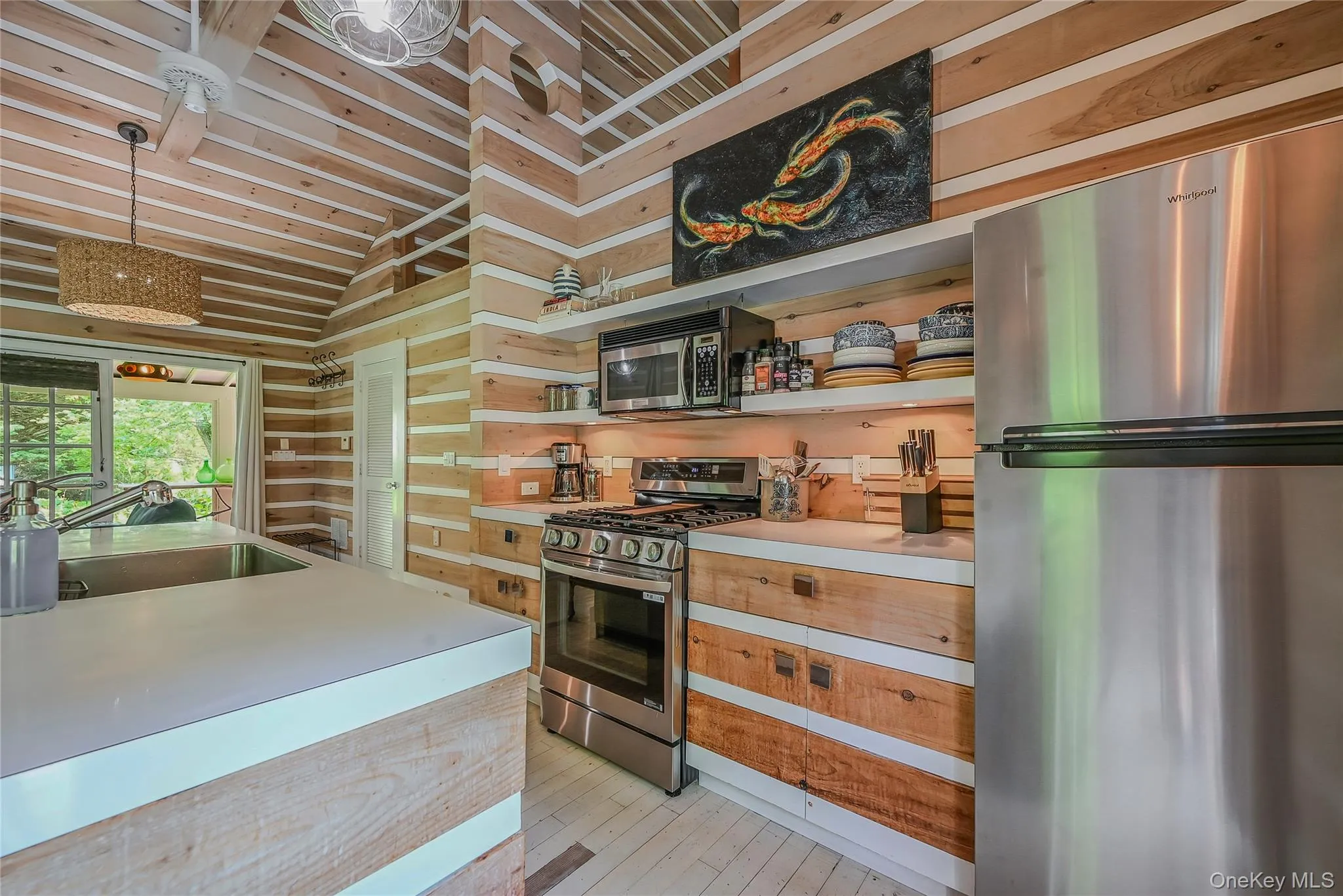 Kitchen with stainless steel appliances, light countertops, open shelves, light wood-style floors, and decorative light fixtures Kitchen with stainless steel appliances, light countertops, open shelves, light wood-style floors, and decorative light fixtures