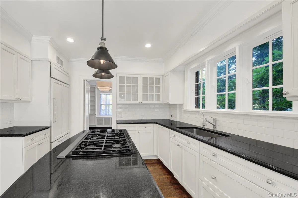 Kitchen featuring an island with a built-in gas cooktop, dark countertops, white cabinetry, a white subway tile backsplash, and multiple windows with grid patterns Kitchen featuring an island with a built-in gas cooktop, dark countertops, white cabinetry, a white subway tile backsplash, and multiple windows with grid patterns