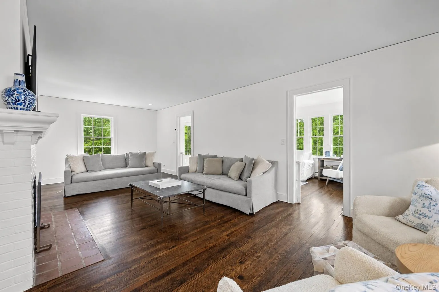Spacious living area featuring rich wood-finish flooring, a painted brick fireplace with a mantel, and bright white walls Spacious living area featuring rich wood-finish flooring, a painted brick fireplace with a mantel, and bright white walls