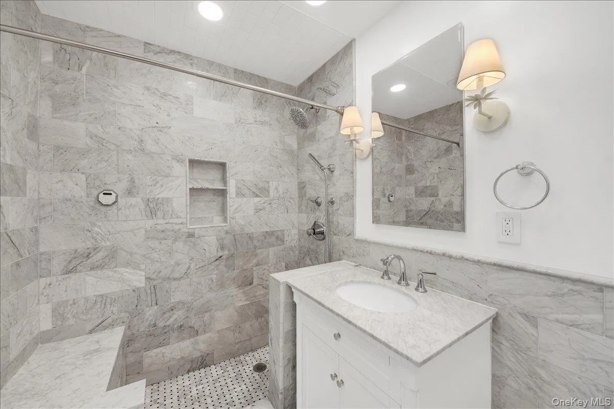 Primary bathroom featuring a tiled shower with integrated bench and niche, a single vanity with a stone countertop, an undermount sink, and wall-mounted sconces Primary bathroom featuring a tiled shower with integrated bench and niche, a single vanity with a stone countertop, an undermount sink, and wall-mounted sconces