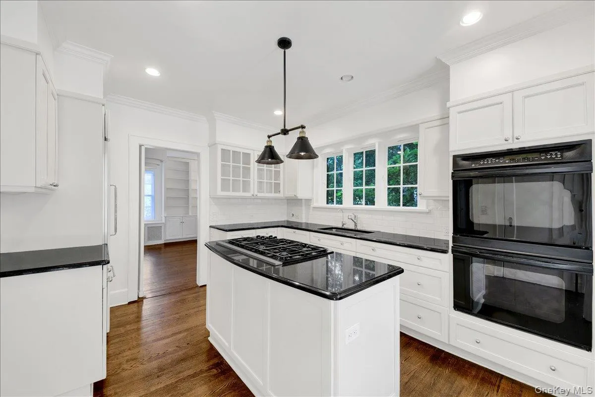 Bright kitchen featuring white cabinetry, dark countertops, hardwood flooring, a gas cooktop on the island, and built-in double ovens Bright kitchen featuring white cabinetry, dark countertops, hardwood flooring, a gas cooktop on the island, and built-in double ovens