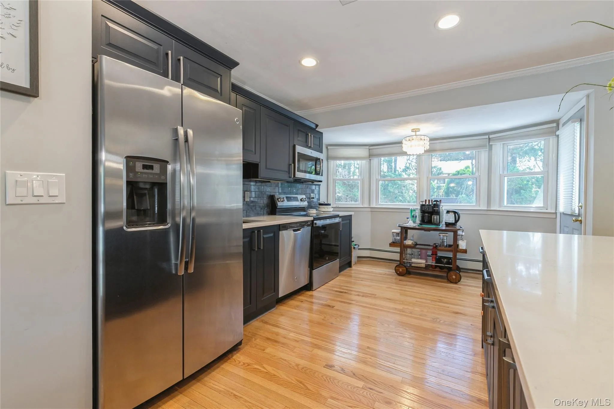 Kitchen featuring stainless steel appliances, dark cabinetry, light countertops, wood-finish flooring, and a bay window configuration with multiple windows Kitchen featuring stainless steel appliances, dark cabinetry, light countertops, wood-finish flooring, and a bay window configuration with multiple windows