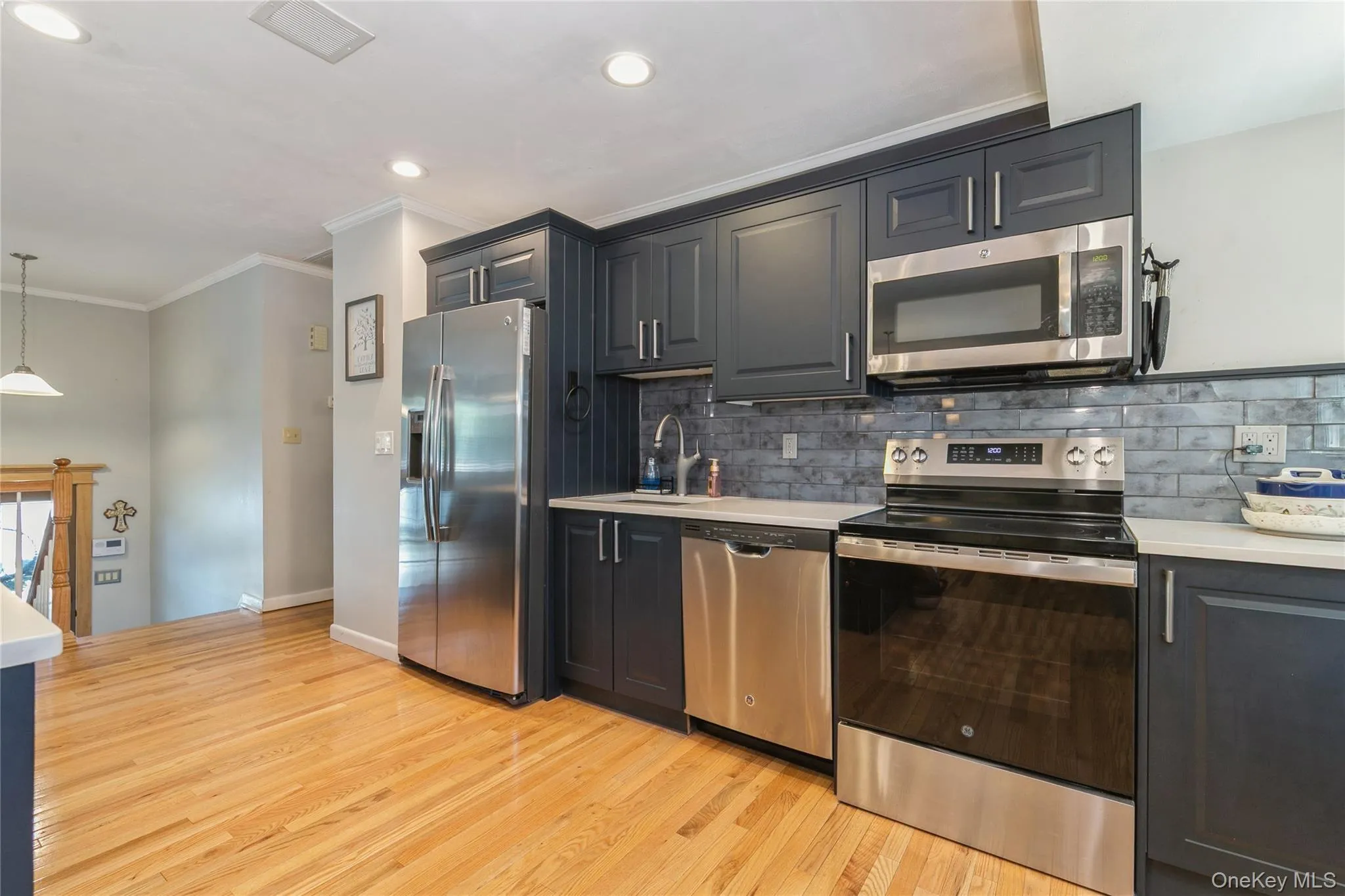 Modern kitchen featuring wood-finish flooring, dark cabinetry, stainless steel appliances, light countertops, and a subway tile backsplash Modern kitchen featuring wood-finish flooring, dark cabinetry, stainless steel appliances, light countertops, and a subway tile backsplash