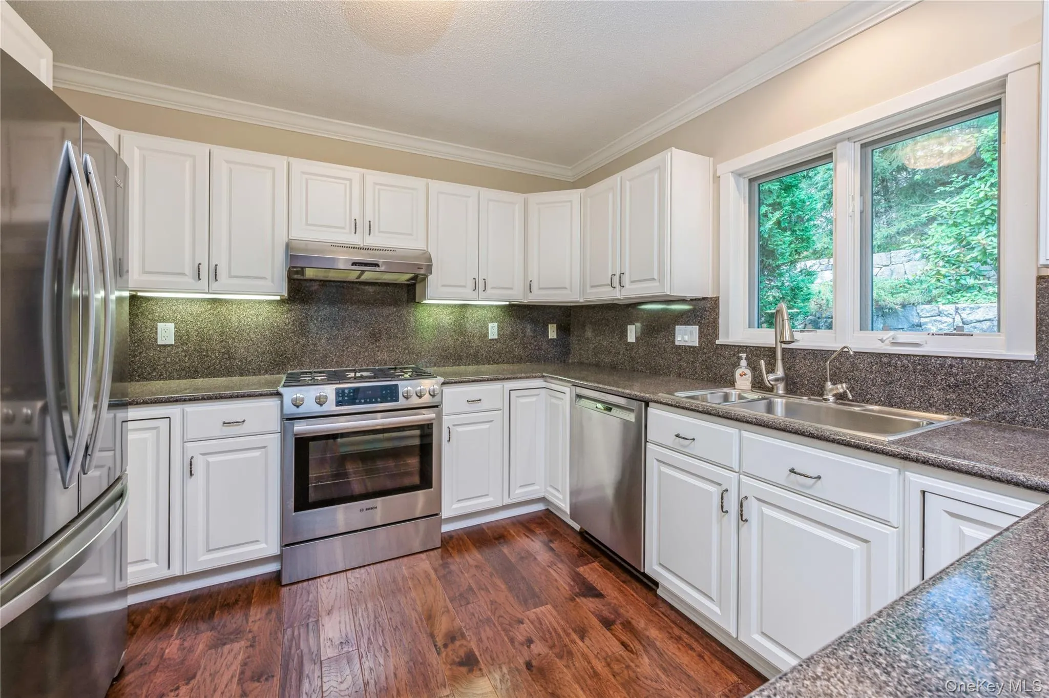 Kitchen with appliances with stainless steel finishes, under cabinet range hood, crown molding, dark countertops, and white cabinetry Kitchen with appliances with stainless steel finishes, under cabinet range hood, crown molding, dark countertops, and white cabinetry