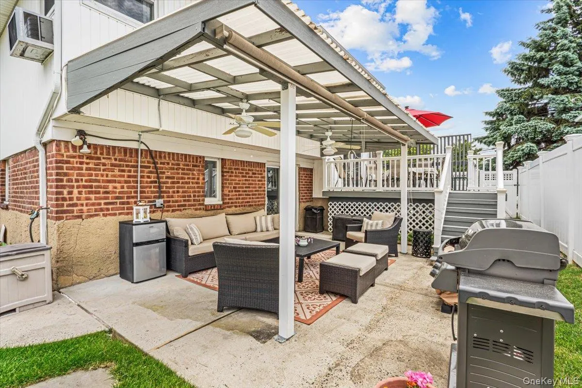 Expansive concrete patio featuring a covered awning with ceiling fans, brick exterior, white vinyl fencing, and a multi-level wooden deck with lattice accents Expansive concrete patio featuring a covered awning with ceiling fans, brick exterior, white vinyl fencing, and a multi-level wooden deck with lattice accents
