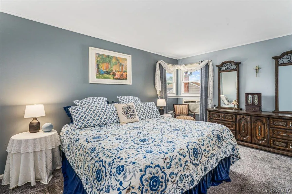 Primary bedroom featuring wall-to-wall carpeting, light blue wall paint, a window with curtains, and a built-in air conditioning unit Primary bedroom featuring wall-to-wall carpeting, light blue wall paint, a window with curtains, and a built-in air conditioning unit