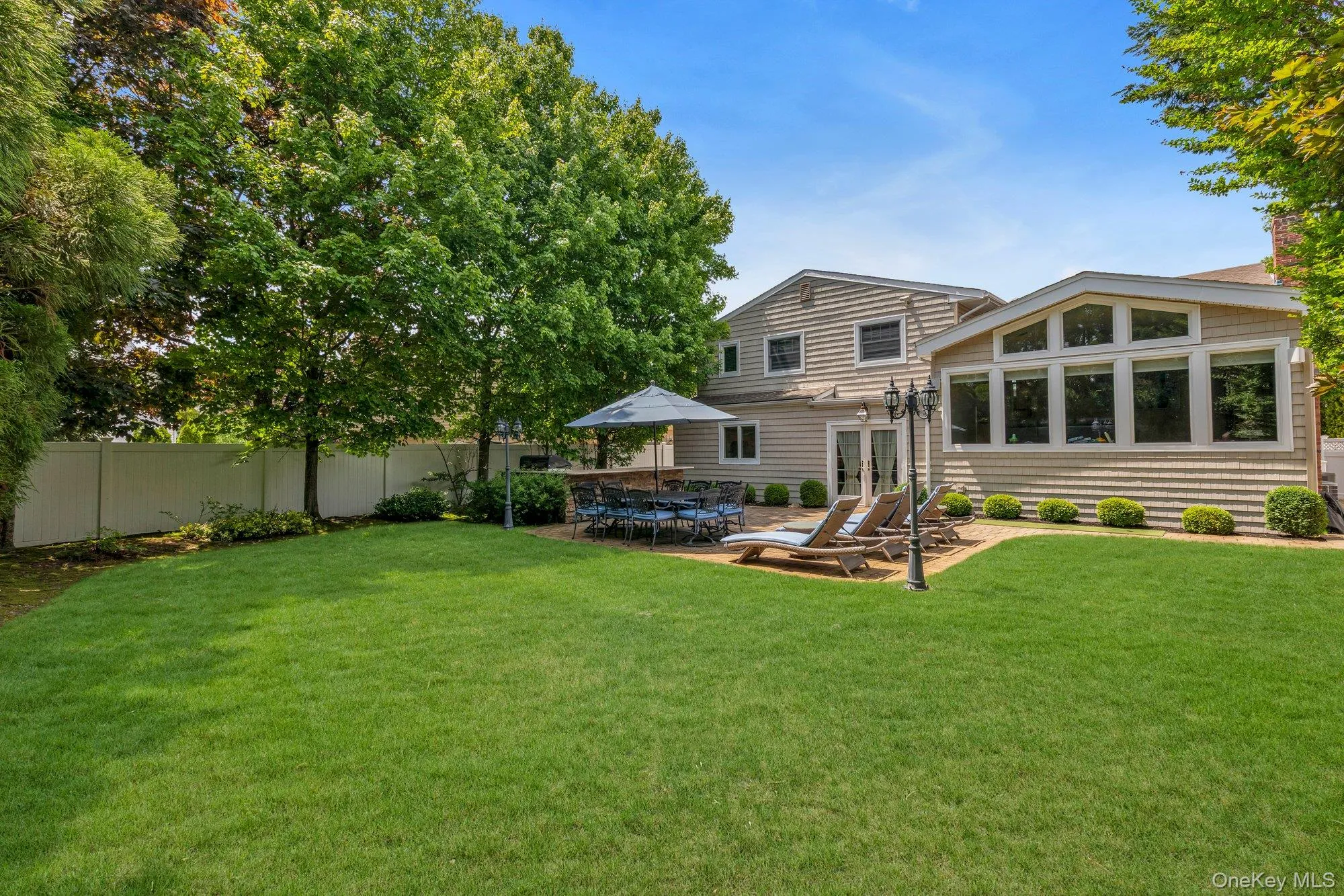 Expansive backyard featuring a large green lawn, paver patio, sunroom addition with a wall of windows, and mature trees providing natural screening Expansive backyard featuring a large green lawn, paver patio, sunroom addition with a wall of windows, and mature trees providing natural screening