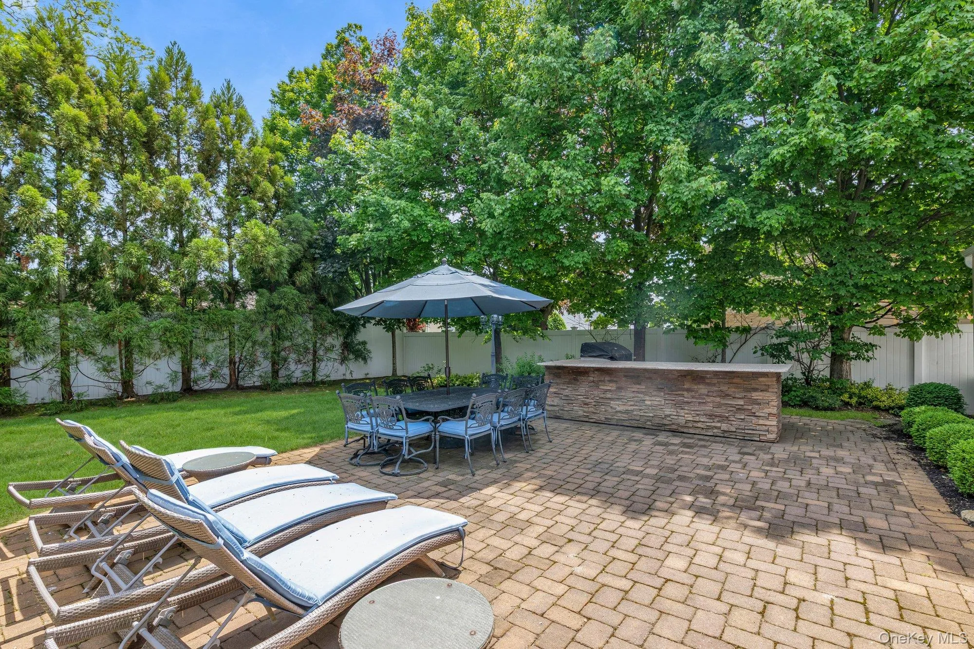 Expansive outdoor living area featuring a paver patio, built-in outdoor kitchen, dedicated dining space, and lush perimeter landscaping Expansive outdoor living area featuring a paver patio, built-in outdoor kitchen, dedicated dining space, and lush perimeter landscaping