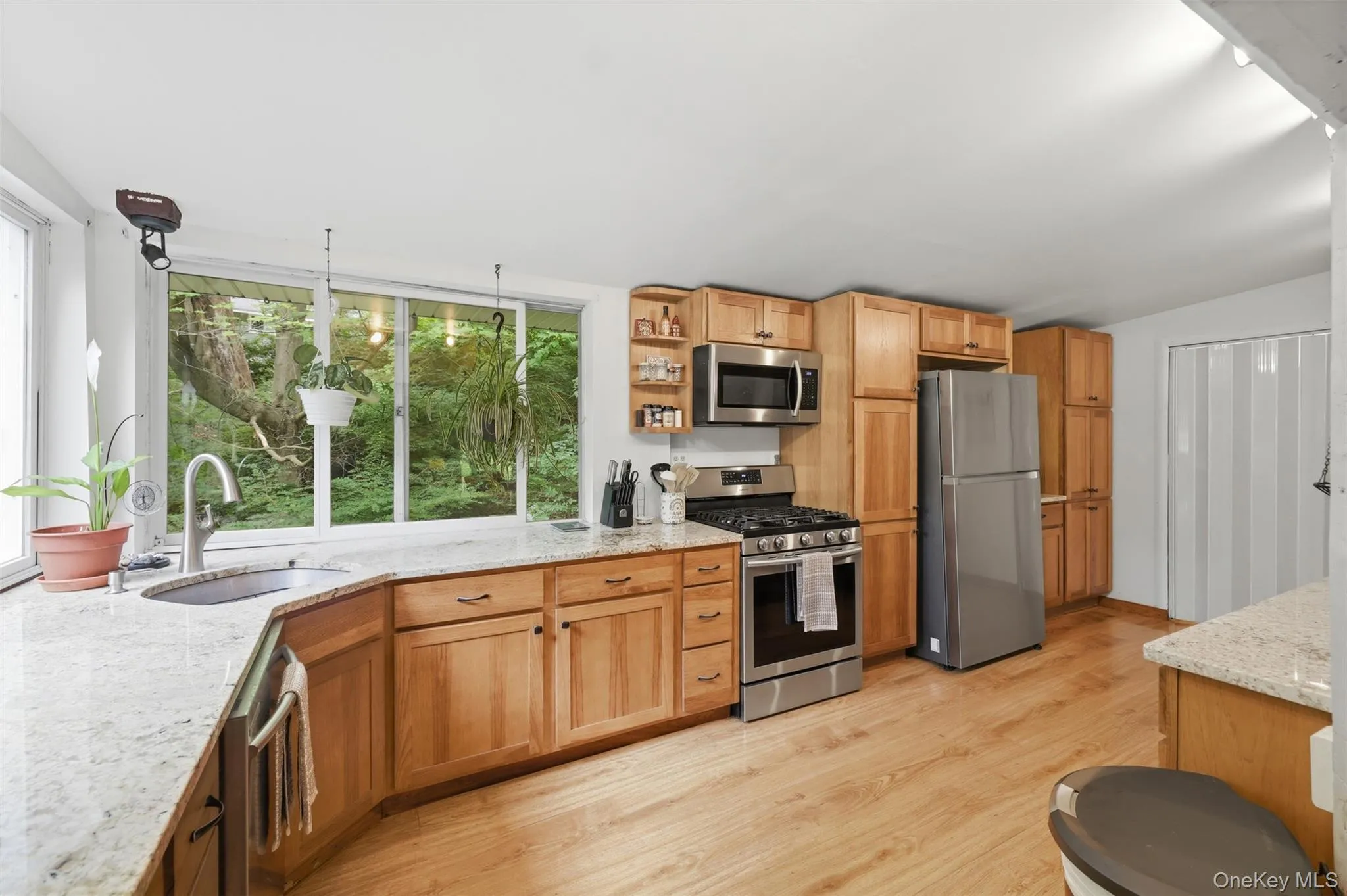 Kitchen with backyard views, featuring light wood cabinetry, light-toned granite countertops, stainless steel appliances, and wood-finish flooring with pantry/laundry through door Kitchen with backyard views, featuring light wood cabinetry, light-toned granite countertops, stainless steel appliances, and wood-finish flooring with pantry/laundry through door