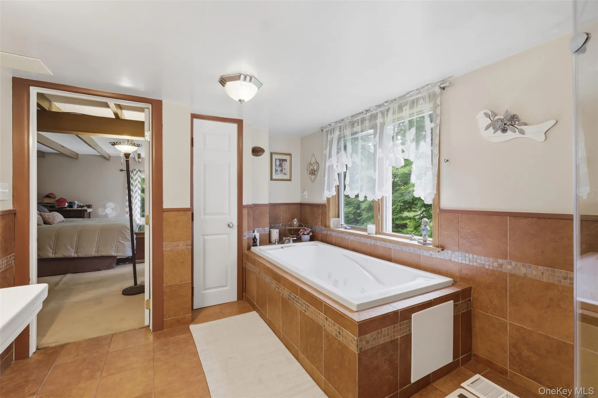 En suite with linen closet and soaking tub surrounded by earth-toned tile, featuring a decorative mosaic border and a window providing natural illumination En suite with linen closet and soaking tub surrounded by earth-toned tile, featuring a decorative mosaic border and a window providing natural illumination