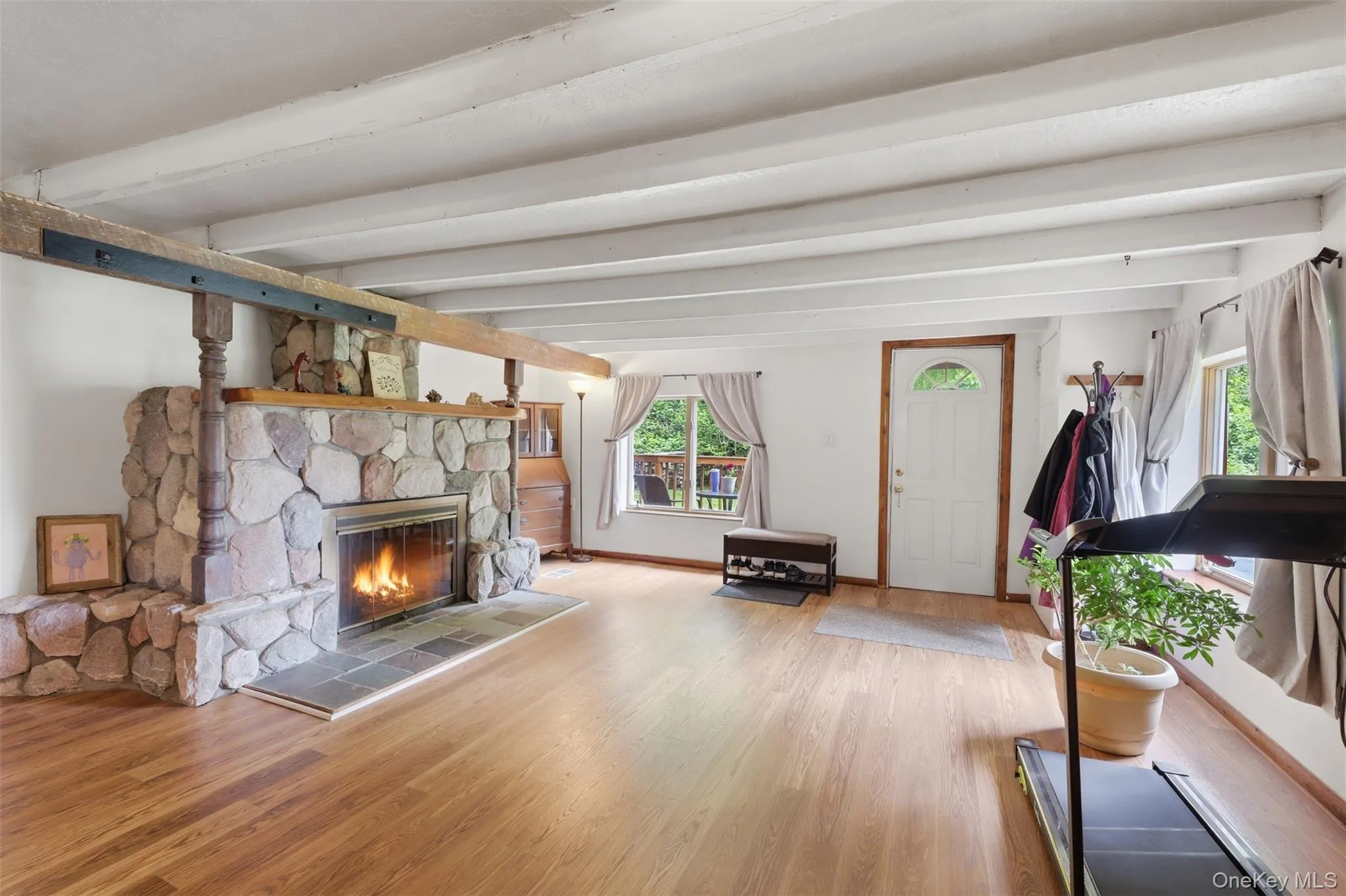 Living area featuring a stone fireplace with a wood mantle, wood-finish flooring, exposed white ceiling beams, and multiple windows, AI-generated flames Living area featuring a stone fireplace with a wood mantle, wood-finish flooring, exposed white ceiling beams, and multiple windows, AI-generated flames