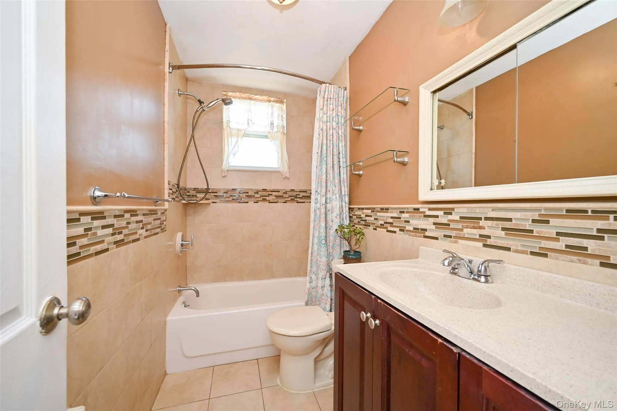 Full bathroom featuring a single vanity with a solid surface countertop, medicine cabinet, tiled bathtub surround with a mosaic accent band, and tiled flooring. Full bathroom featuring a single vanity with a solid surface countertop, medicine cabinet, tiled bathtub surround with a mosaic accent band, and tiled flooring.