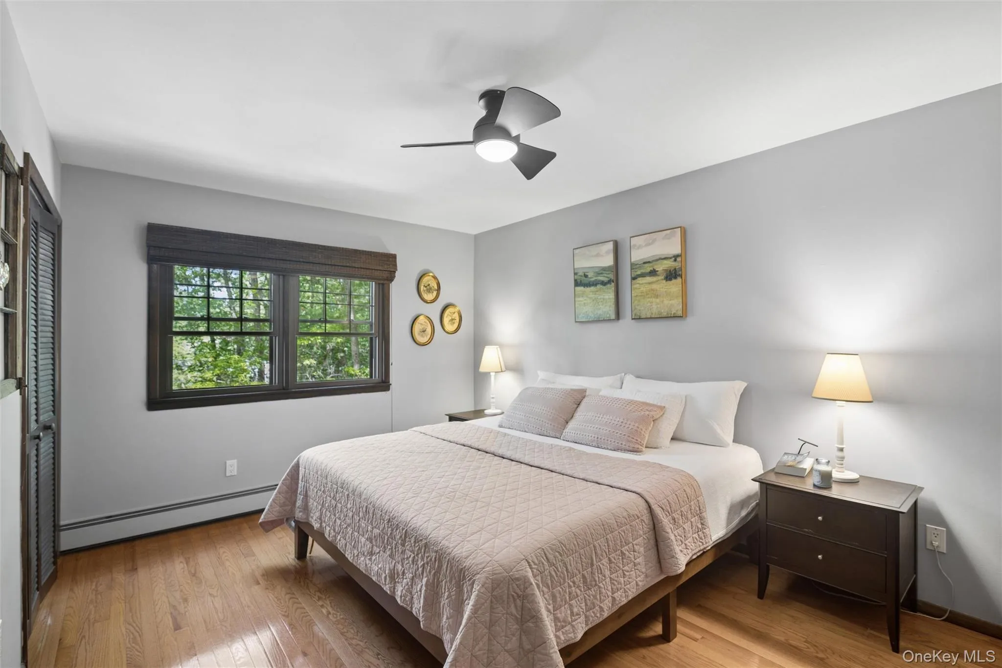 Bedroom featuring wood-finish flooring, a ceiling fan, a window with dark trim, and a built-in louvered door Bedroom featuring wood-finish flooring, a ceiling fan, a window with dark trim, and a built-in louvered door