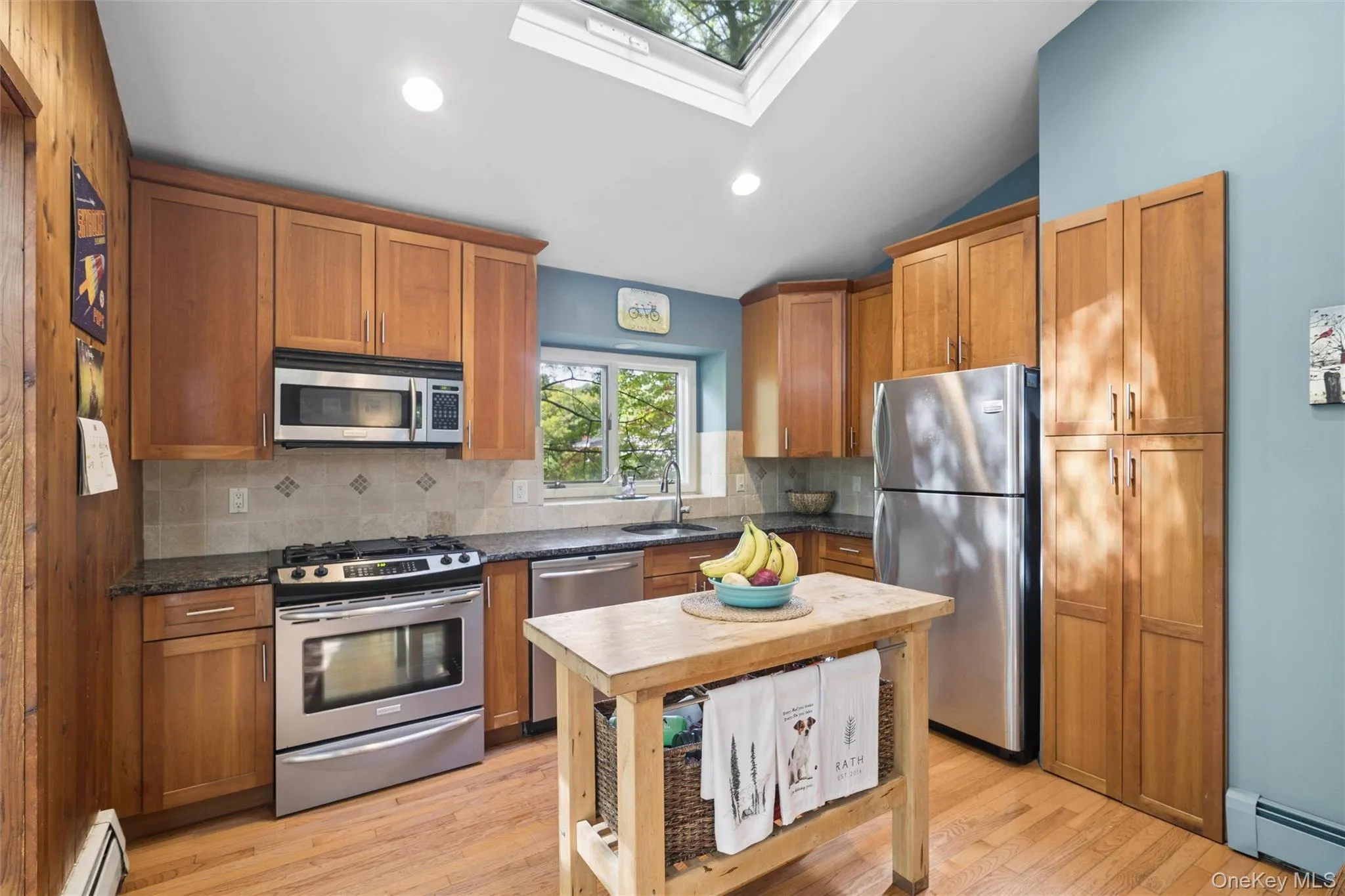 Kitchen featuring a skylight, wood-finish cabinetry, stainless steel appliances, dark countertops, and a tile backsplash Kitchen featuring a skylight, wood-finish cabinetry, stainless steel appliances, dark countertops, and a tile backsplash