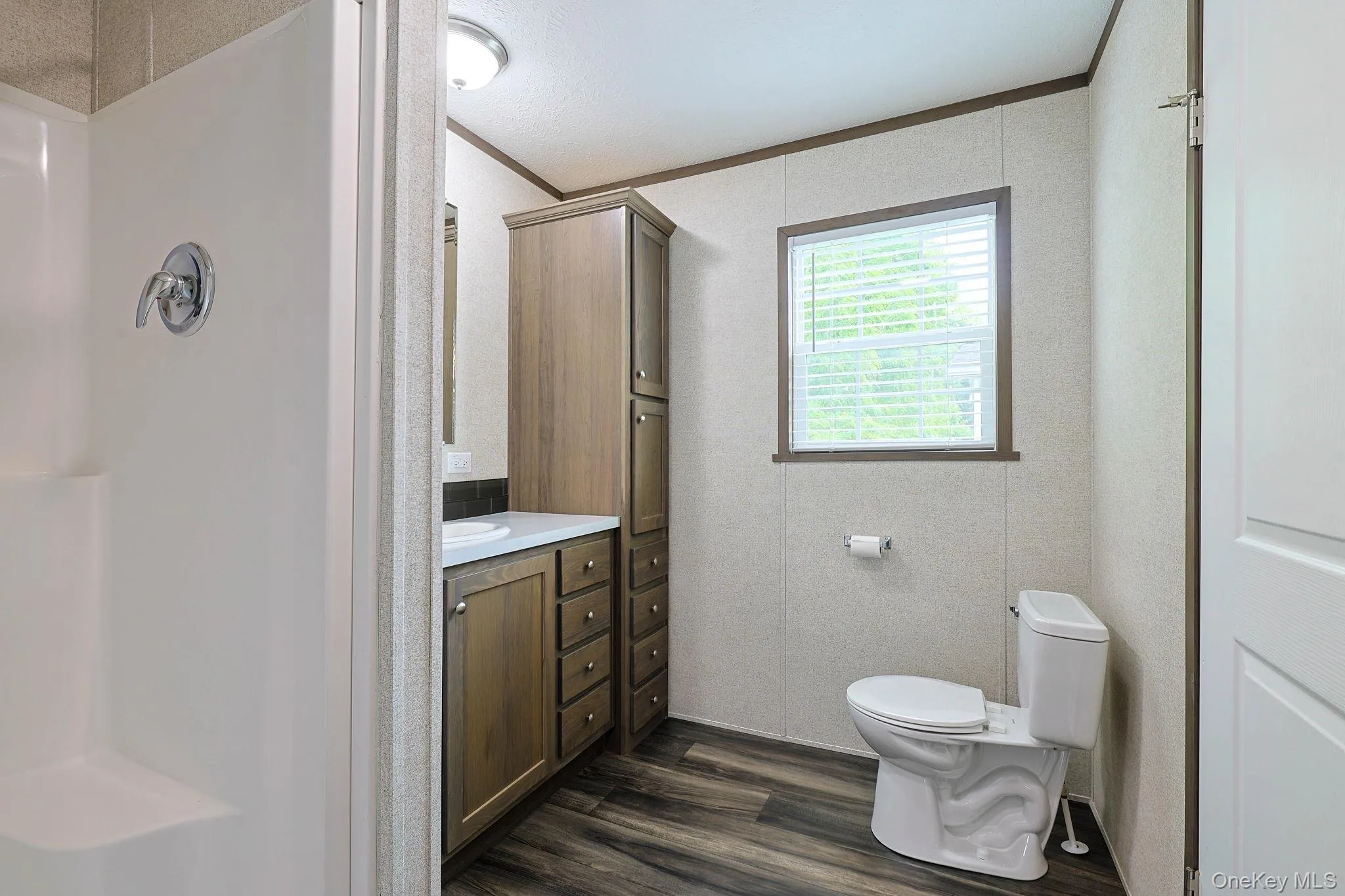 Bathroom featuring a white shower stall, wood-finish flooring, a vanity with a white countertop and wood-tone cabinetry, a tall wood-tone storage cabinet, and a window with blinds Bathroom featuring a white shower stall, wood-finish flooring, a vanity with a white countertop and wood-tone cabinetry, a tall wood-tone storage cabinet, and a window with blinds