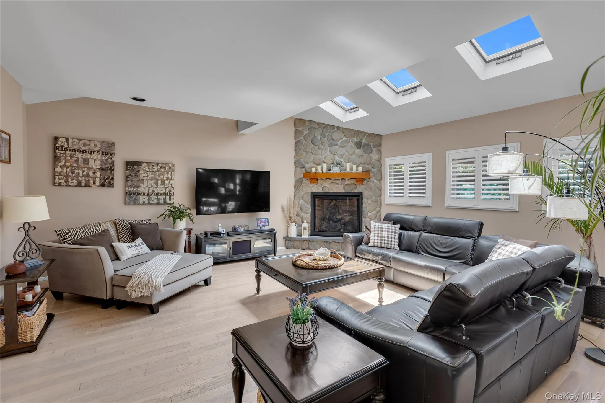 Living area featuring an angled ceiling with multiple skylights, a stone fireplace with a wood mantle, and light wood-finish flooring Living area featuring an angled ceiling with multiple skylights, a stone fireplace with a wood mantle, and light wood-finish flooring