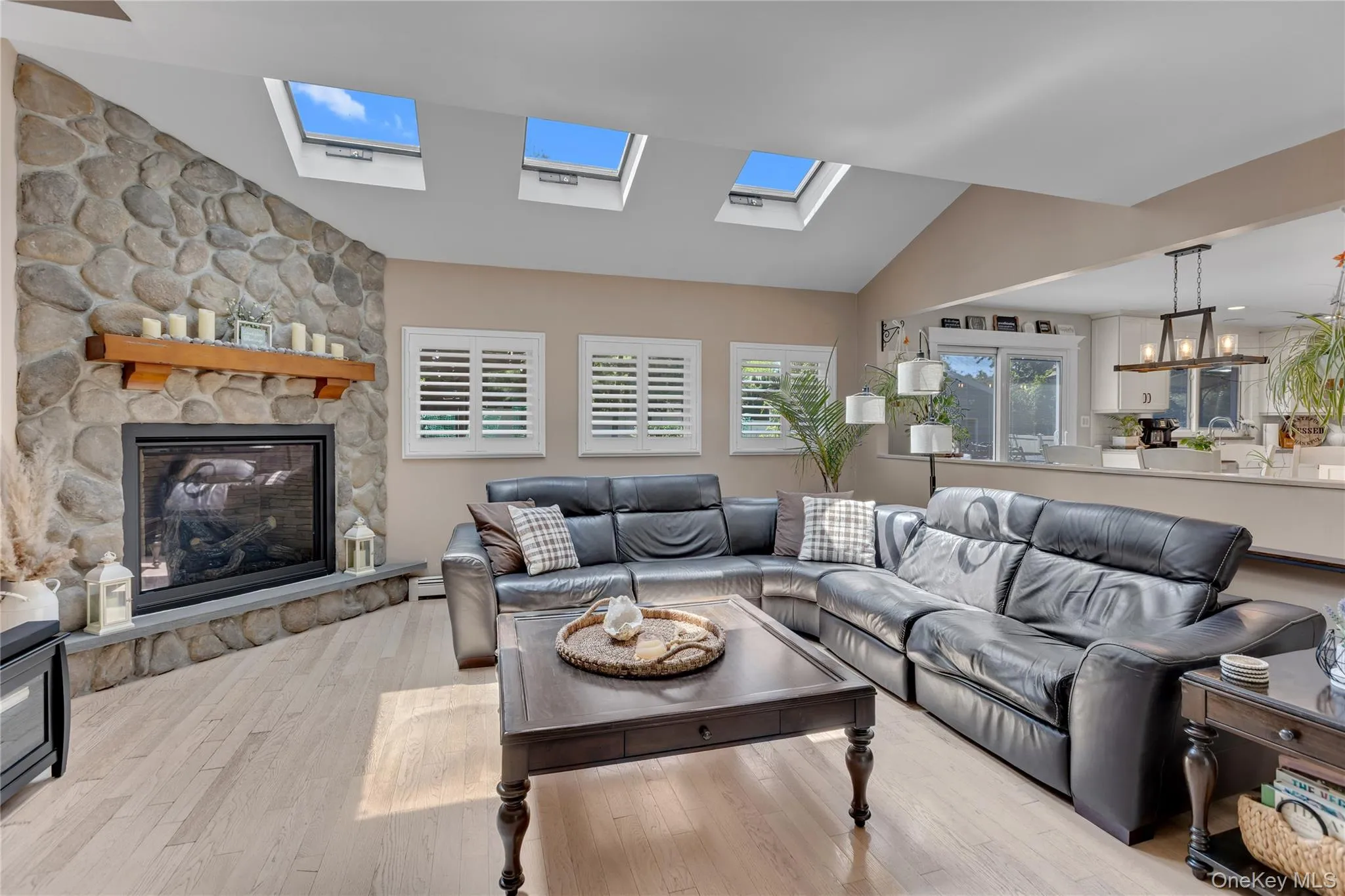 Vaulted ceiling with multiple skylights, stone-clad fireplace with a wood mantle, wood-finish flooring, two windows with white plantation shutters, and a kitchen pass-through Vaulted ceiling with multiple skylights, stone-clad fireplace with a wood mantle, wood-finish flooring, two windows with white plantation shutters, and a kitchen pass-through