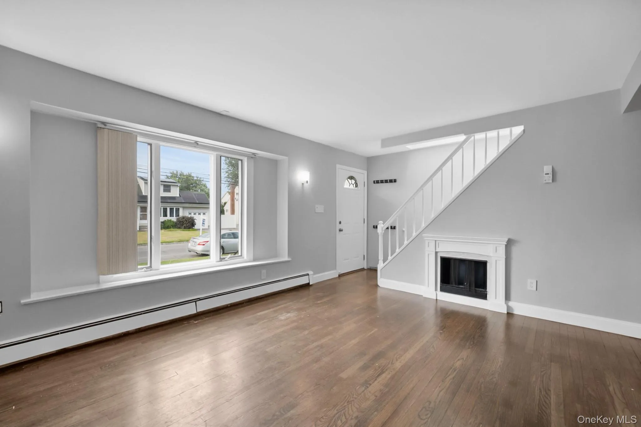 Spacious living area featuring rich wood-finish flooring, a prominent front window with a deep sill, a decorative fireplace with white mantel, a white staircase with balusters, and a white entry door with an arched glass insert Spacious living area featuring rich wood-finish flooring, a prominent front window with a deep sill, a decorative fireplace with white mantel, a white staircase with balusters, and a white entry door with an arched glass insert