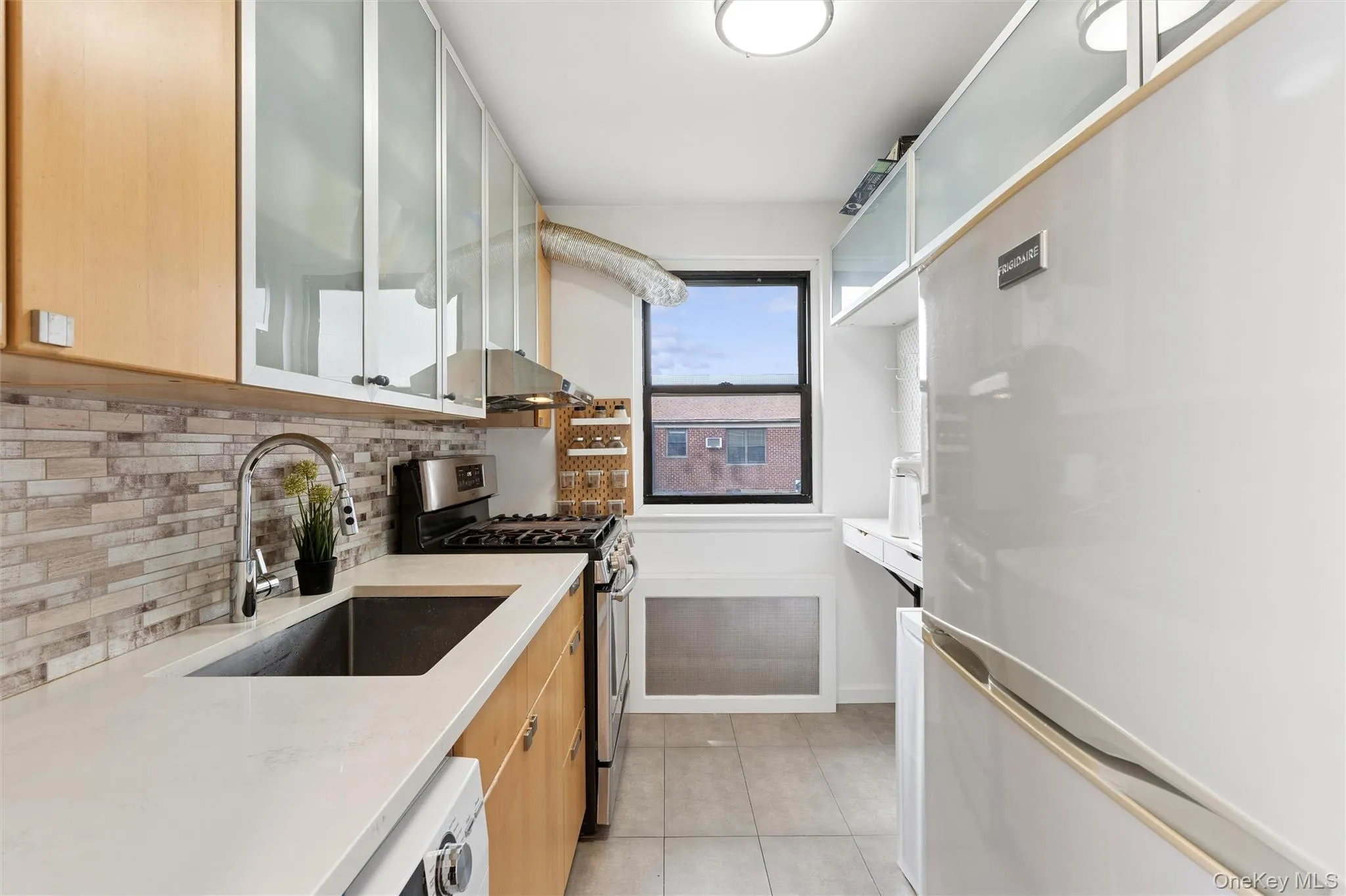 Galley kitchen featuring light wood-finish cabinetry with frosted glass upper doors, stainless steel appliances, and a mosaic tile backsplash Galley kitchen featuring light wood-finish cabinetry with frosted glass upper doors, stainless steel appliances, and a mosaic tile backsplash
