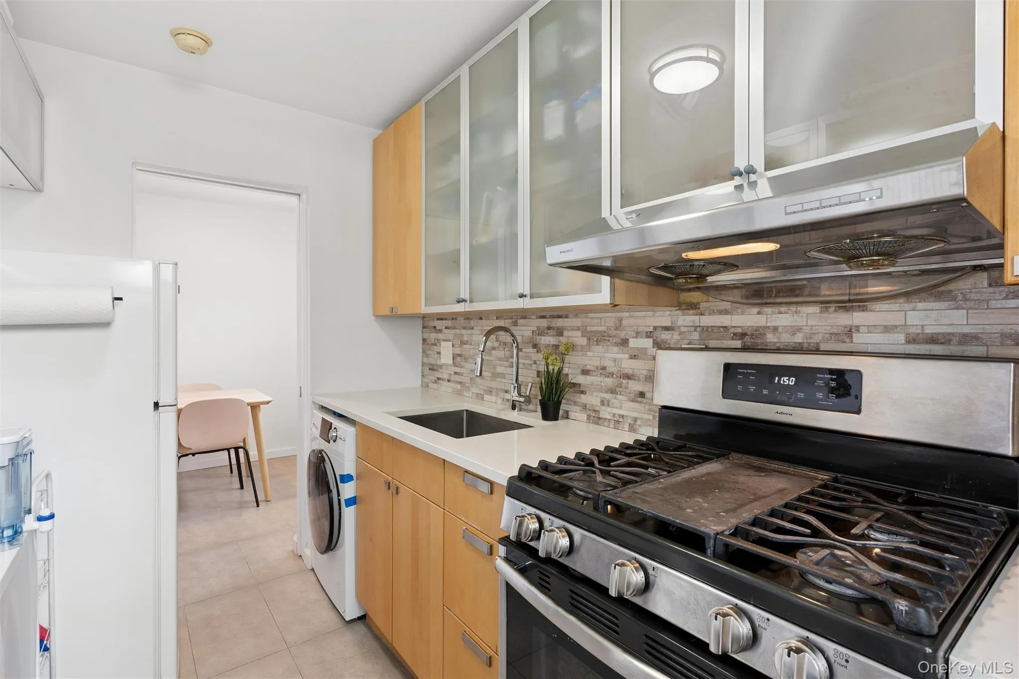 Kitchen featuring a gas range with a stainless steel hood, light wood-finish cabinetry with frosted glass upper doors, a white countertop, and a stacked stone backsplash Kitchen featuring a gas range with a stainless steel hood, light wood-finish cabinetry with frosted glass upper doors, a white countertop, and a stacked stone backsplash