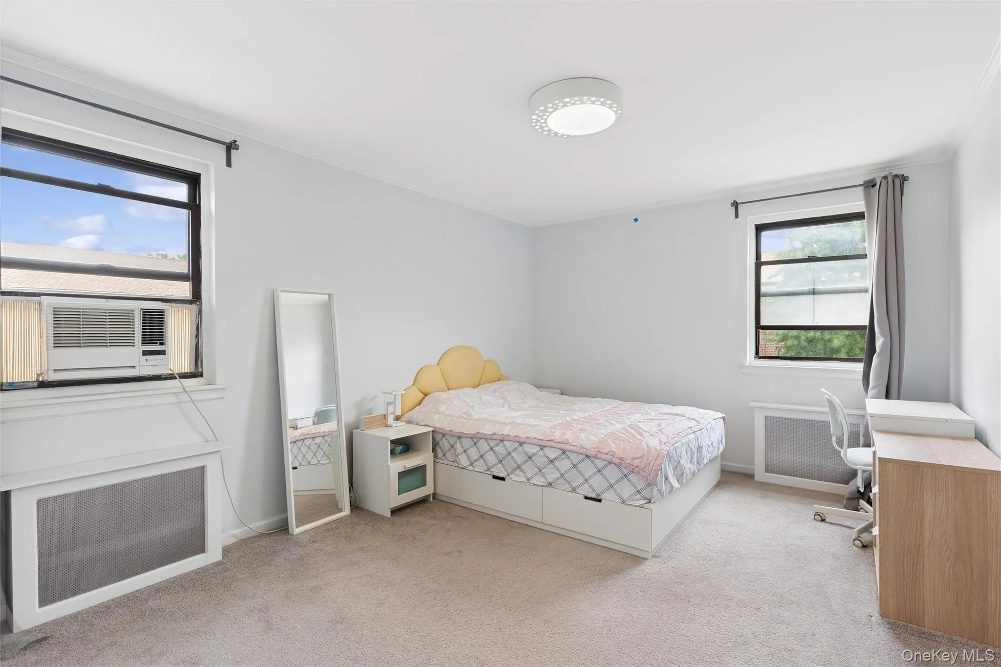 Spacious primary bedroom featuring two large windows, light gray wall paint, and gray carpeted flooring Spacious primary bedroom featuring two large windows, light gray wall paint, and gray carpeted flooring