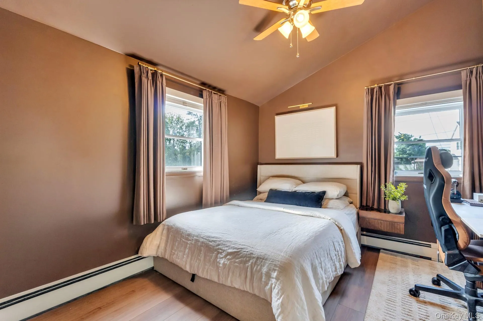 Vaulted ceiling bedroom featuring wood-finish flooring, two windows with drapery, a ceiling fan, and baseboard heating Vaulted ceiling bedroom featuring wood-finish flooring, two windows with drapery, a ceiling fan, and baseboard heating
