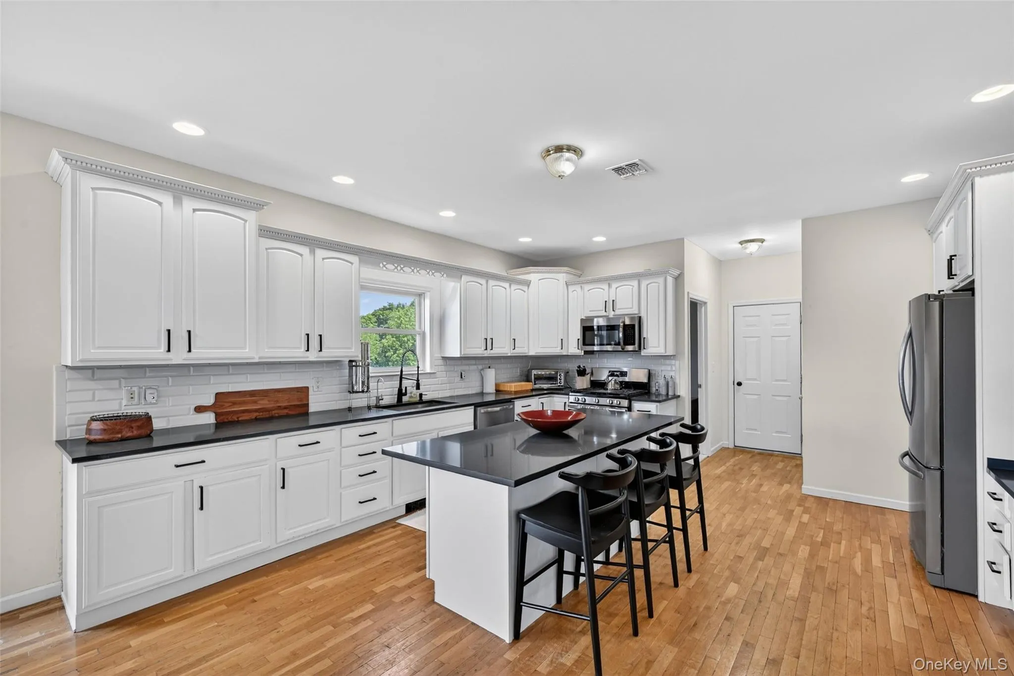 Kitchen featuring a central island with countertop seating, white shaker-style cabinetry with crown molding, dark countertops, subway tile backsplash, and light wood-finish flooring Kitchen featuring a central island with countertop seating, white shaker-style cabinetry with crown molding, dark countertops, subway tile backsplash, and light wood-finish flooring