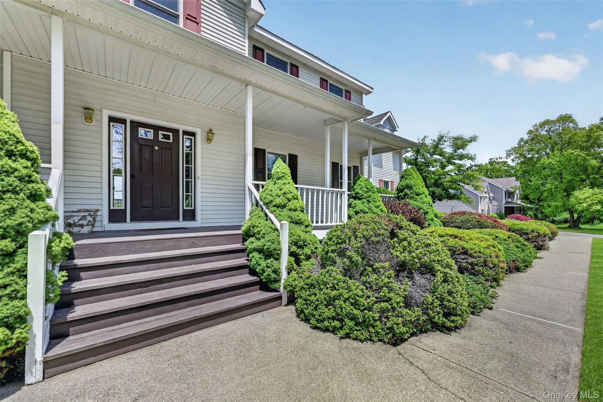 Inviting front porch featuring a dark wood-finish entry door with sidelights, white siding exterior, and well-maintained shrubbery Inviting front porch featuring a dark wood-finish entry door with sidelights, white siding exterior, and well-maintained shrubbery