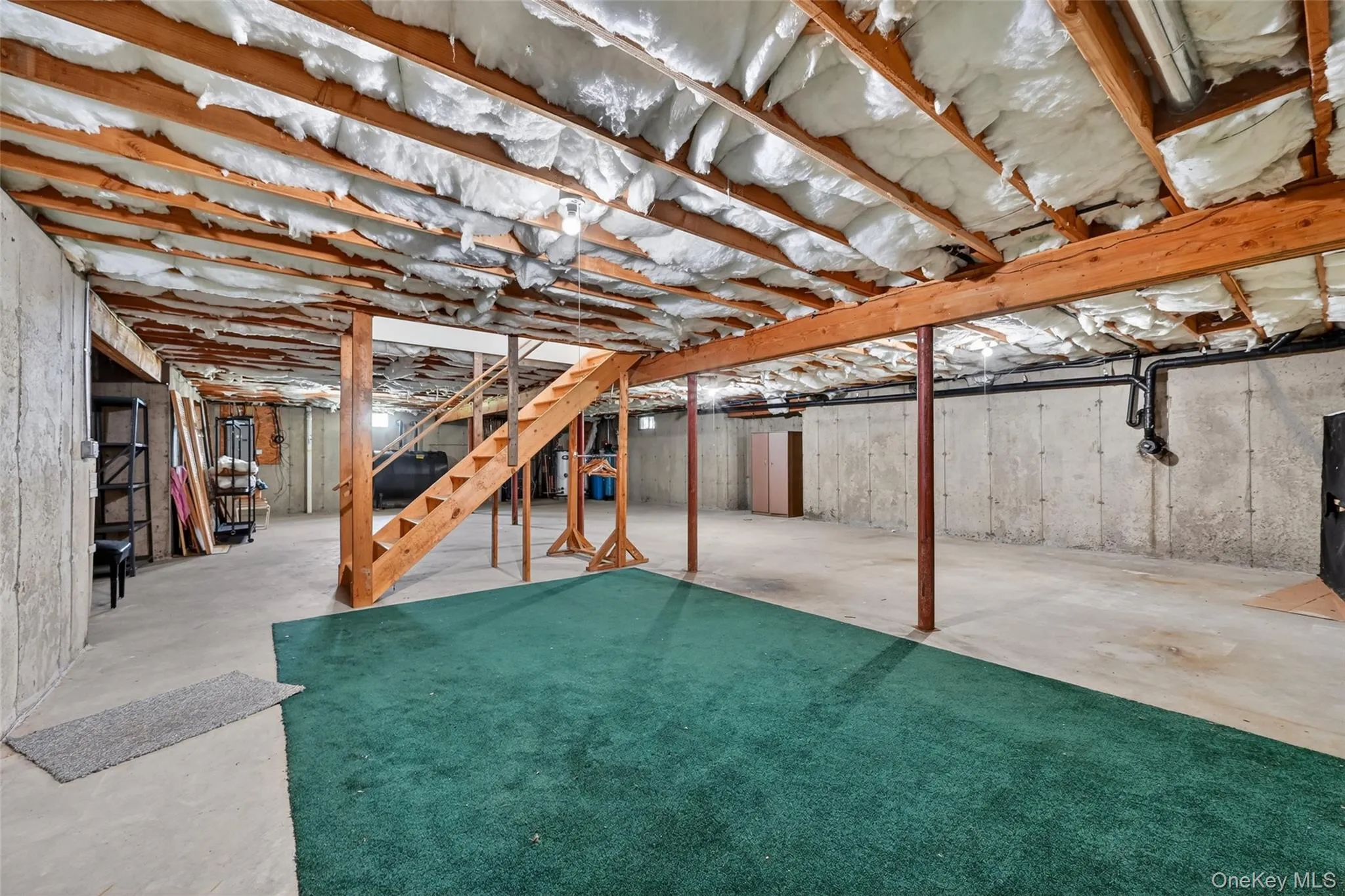 Unfinished basement featuring exposed wood joists with insulation, concrete slab flooring, and a partial green carpet area Unfinished basement featuring exposed wood joists with insulation, concrete slab flooring, and a partial green carpet area