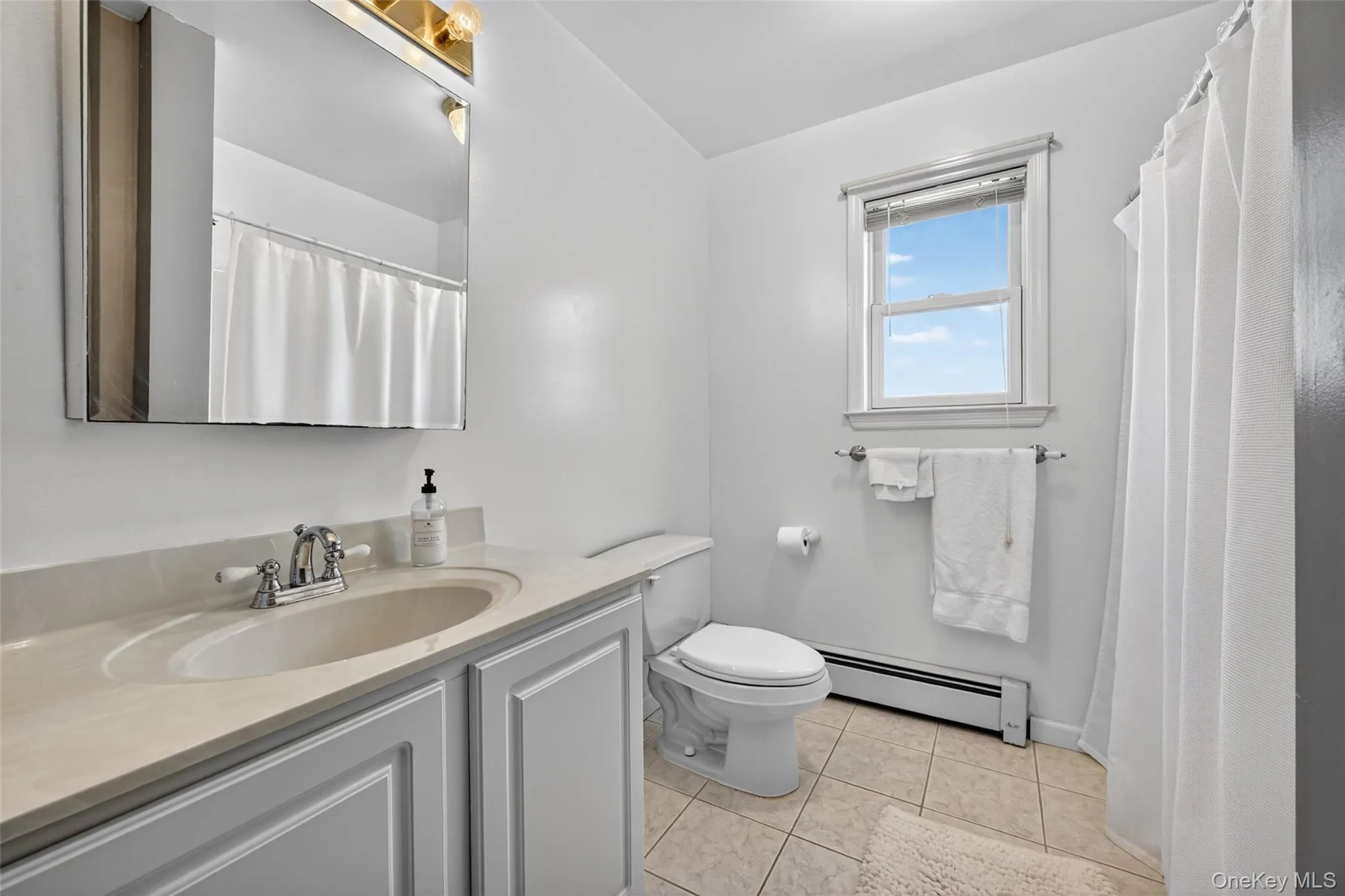 Bathroom featuring a single vanity with a light-toned countertop, a window with blinds, a toilet, a full shower/tub combination with a curtain, and tile flooring Bathroom featuring a single vanity with a light-toned countertop, a window with blinds, a toilet, a full shower/tub combination with a curtain, and tile flooring