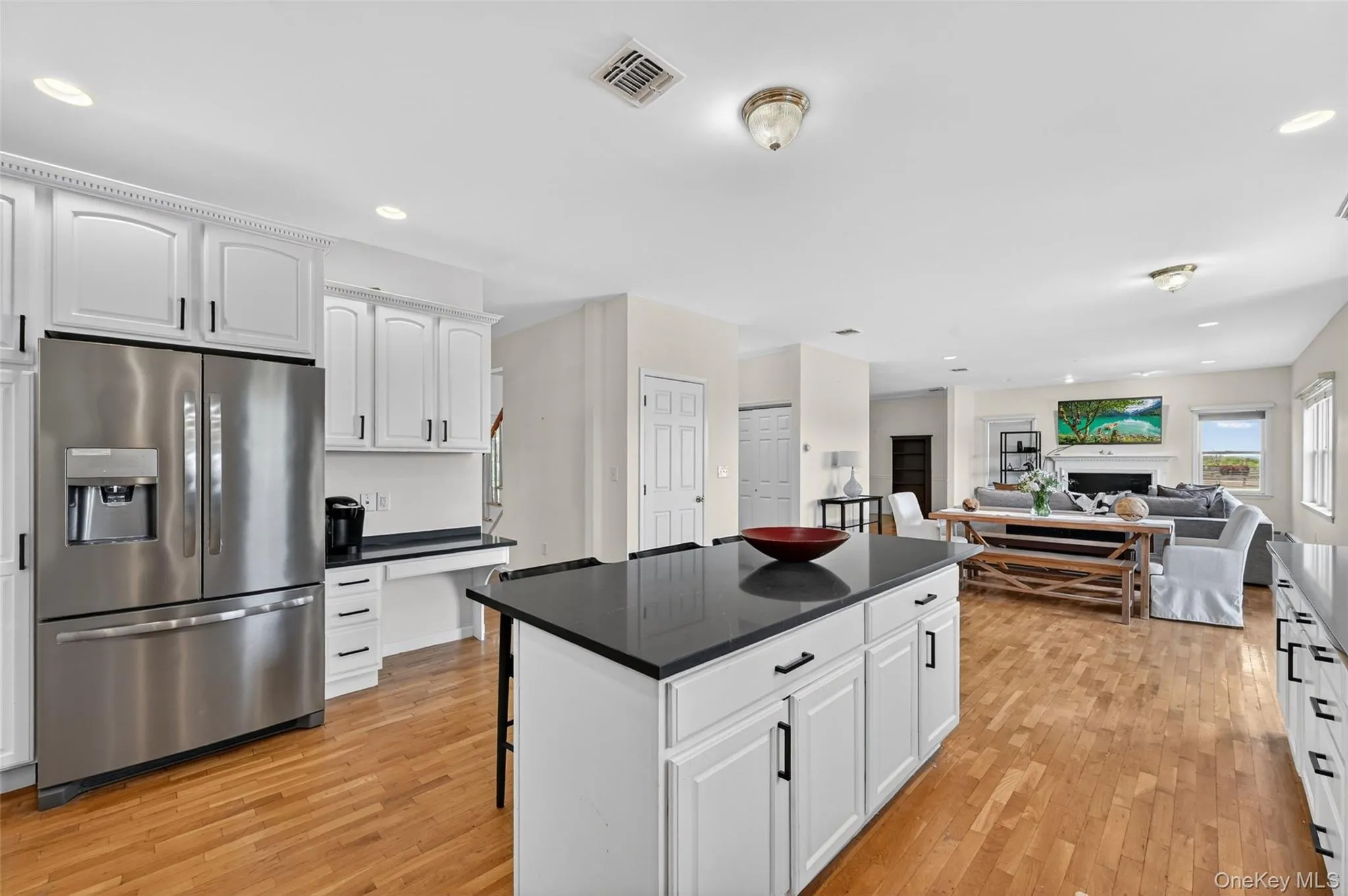 Open-concept kitchen featuring an island with a dark Granite countertop, white cabinetry with black hardware, stainless steel refrigerator, and wood-finish flooring Open-concept kitchen featuring an island with a dark Granite countertop, white cabinetry with black hardware, stainless steel refrigerator, and wood-finish flooring