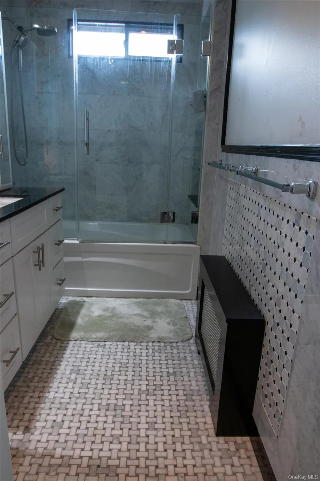 Bathroom featuring a glass-enclosed shower over a tub, white vanity cabinetry with dark countertops, a mosaic tile accent wall, and woven pattern floor tiling Bathroom featuring a glass-enclosed shower over a tub, white vanity cabinetry with dark countertops, a mosaic tile accent wall, and woven pattern floor tiling