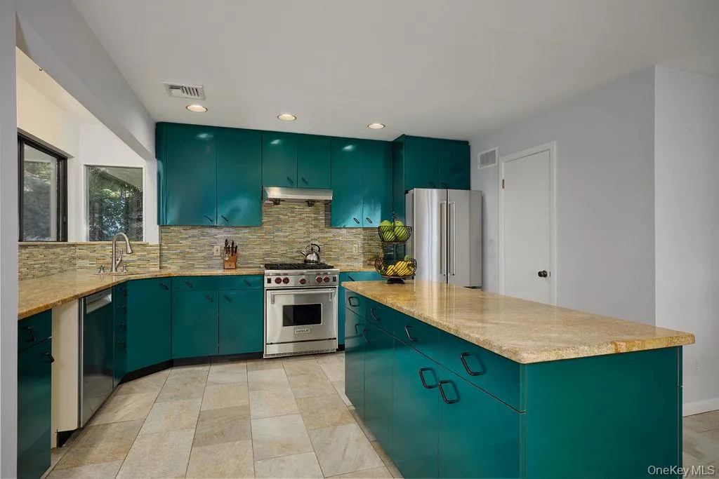 Kitchen featuring teal cabinetry with black hardware, stone-finish countertops, tiled backsplash, stainless steel appliances, and a central island Kitchen featuring teal cabinetry with black hardware, stone-finish countertops, tiled backsplash, stainless steel appliances, and a central island