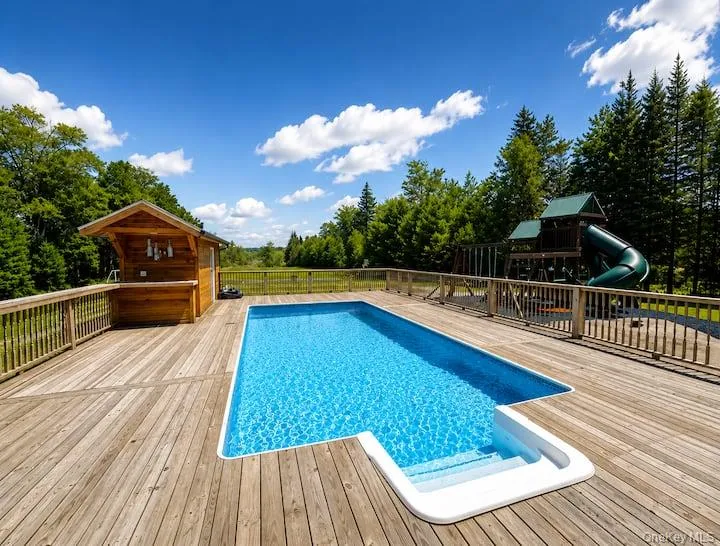 Expansive outdoor deck featuring an in-ground swimming pool with built-in steps, a dedicated poolside bar structure, and surrounding wooded landscape Expansive outdoor deck featuring an in-ground swimming pool with built-in steps, a dedicated poolside bar structure, and surrounding wooded landscape