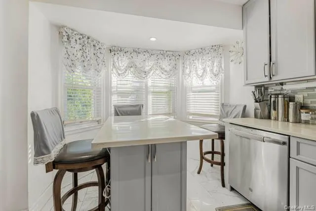 Kitchen area featuring a bay window, recessed lighting, light gray cabinetry, stainless steel dishwasher, and a light-toned countertop Kitchen area featuring a bay window, recessed lighting, light gray cabinetry, stainless steel dishwasher, and a light-toned countertop