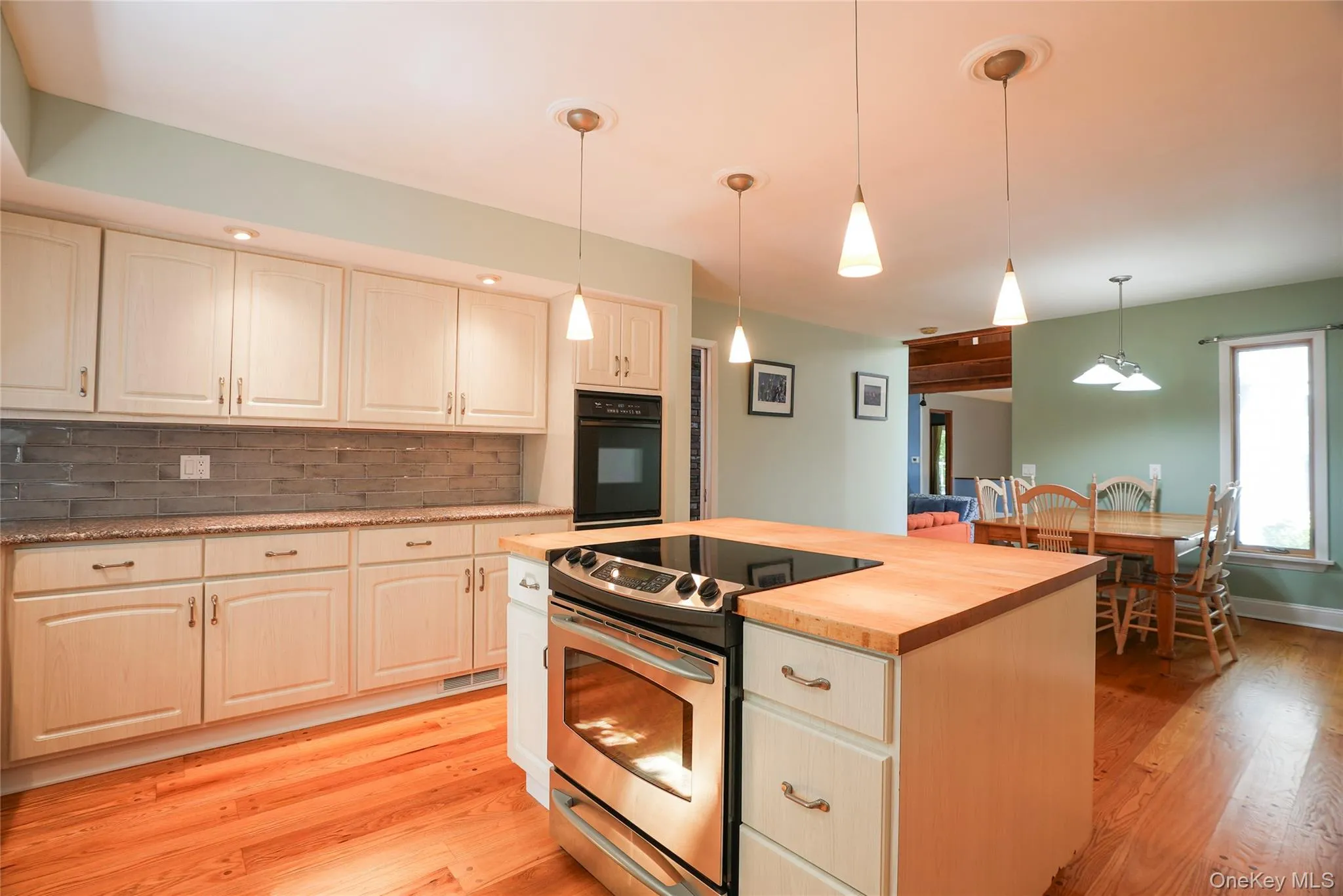 Kitchen featuring a center island with a built-in range, pendant lighting, hardwood flooring, light-toned cabinetry, and a stone-tiled backsplash Kitchen featuring a center island with a built-in range, pendant lighting, hardwood flooring, light-toned cabinetry, and a stone-tiled backsplash